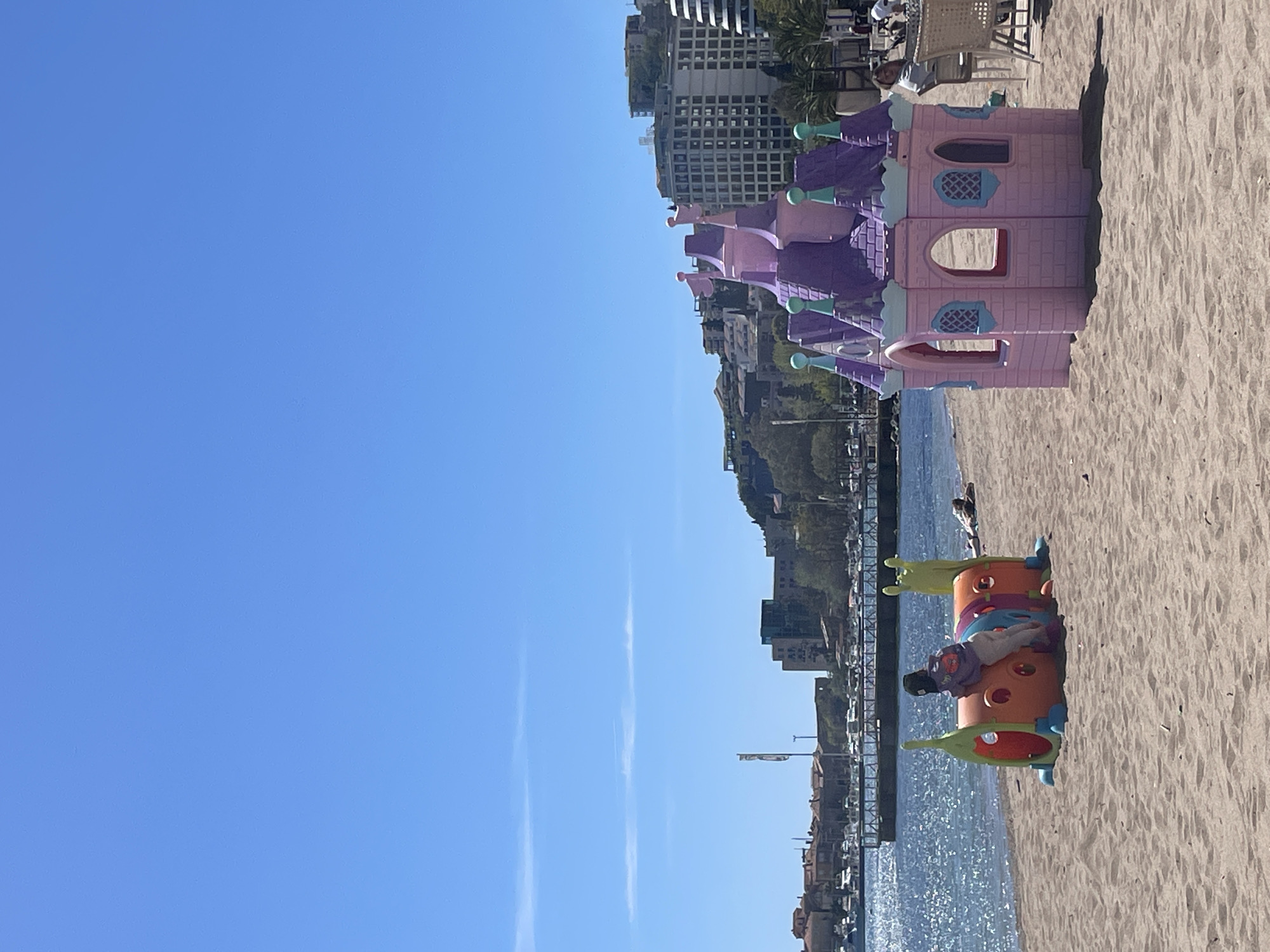 On a sunny beach in Budva, Montenegro, with a clear blue sky, a pink and purple plastic play castle stands on the sand. A child plays in a colorful caterpillar-shaped structure nearby. The Adriatic Sea laps the shore, with buildings lining the coast in the background. Chairs and tables are set up on a patio near the castle.