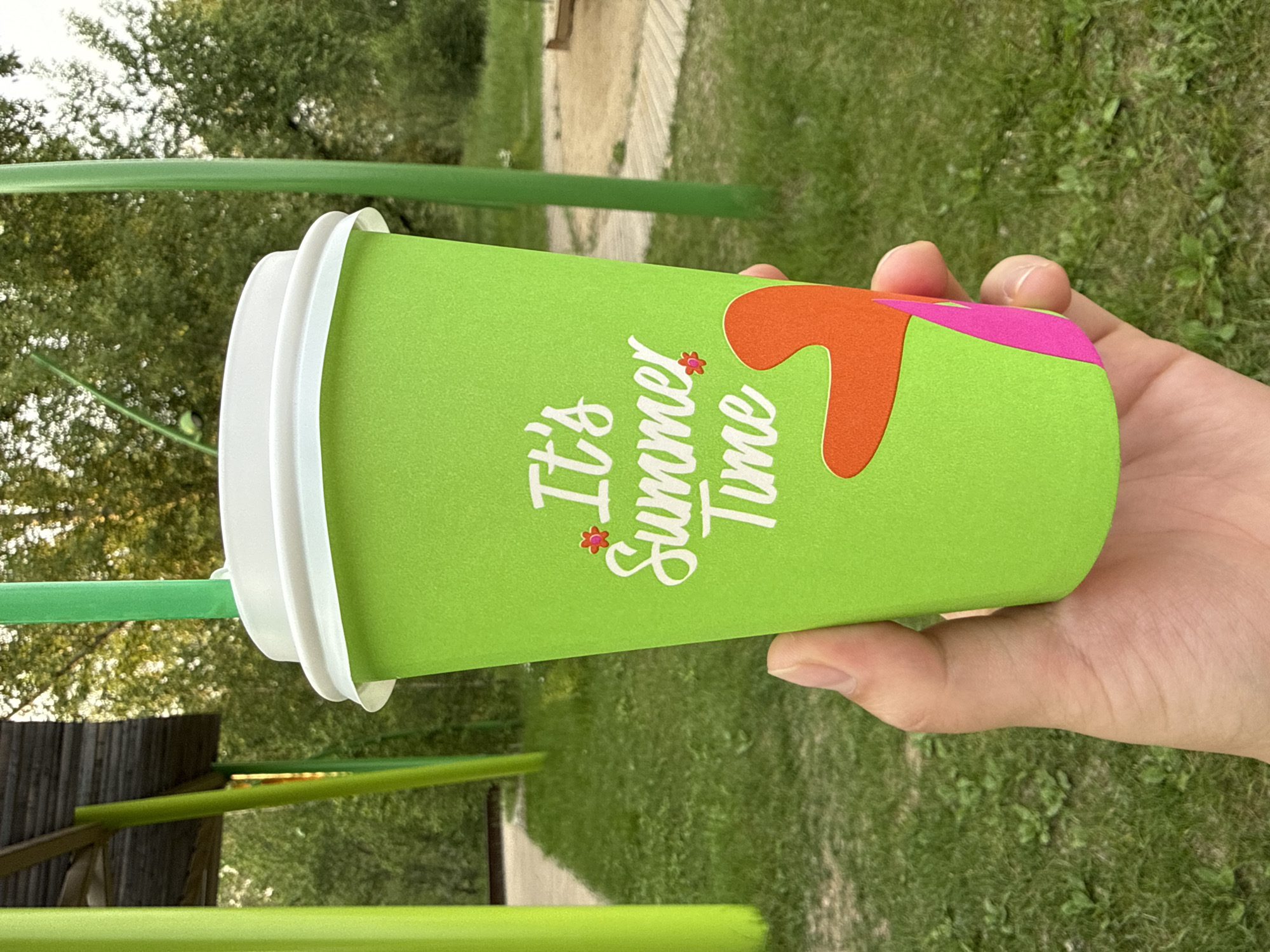A person's hand holds a bright green disposable cup with a white lid and a green straw. The cup features white text that reads "It's Summer Time" and abstract colorful shapes in orange and pink. The background is an outdoor setting with green grass and trees, suggesting a park or recreational area. The image is taken in Krasnoyarsk, Russia.