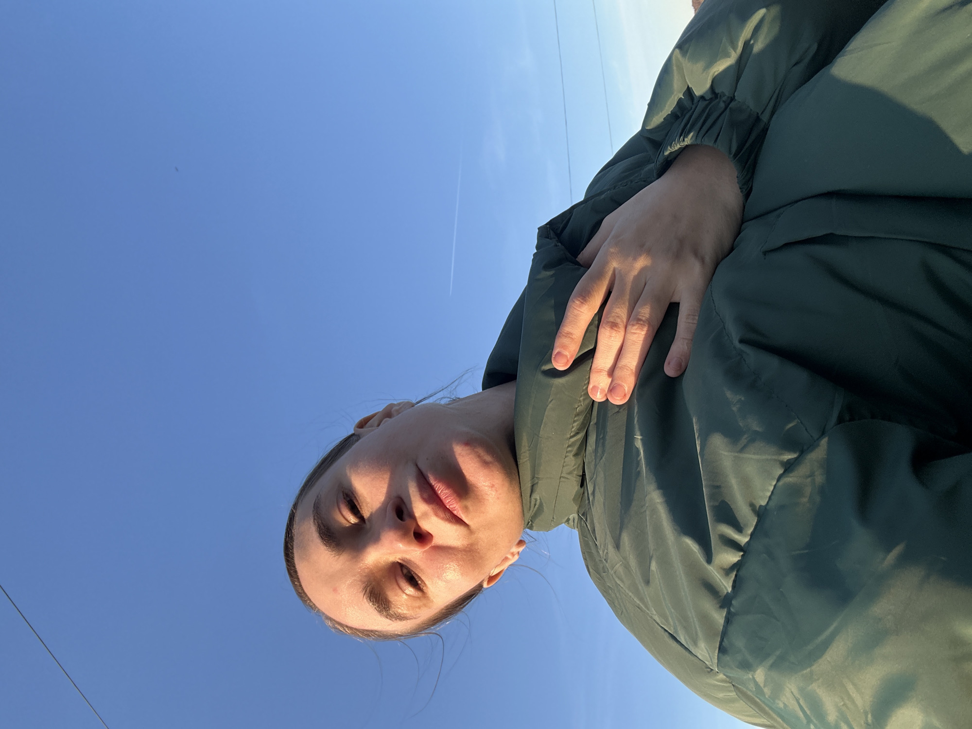A low-angle shot shows a person wearing a dark green puffy jacket against a clear blue sky. The person's face is illuminated by sunlight, with a neutral expression. Power lines stretch across the top-left portion of the sky, and a contrail from an airplane is visible in the distance. The location cannot be determined.