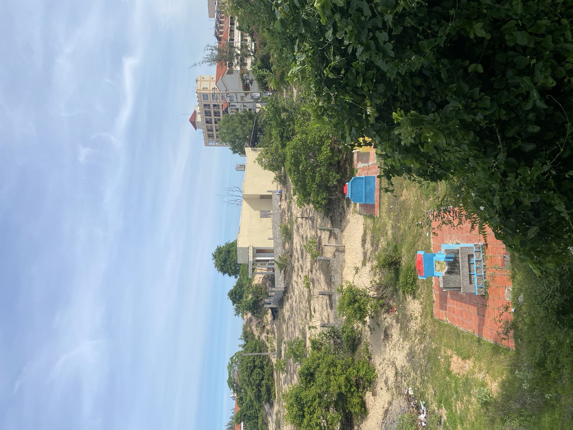 The image depicts a scene in Mũi Né, Vietnam, characterized by a mix of natural elements and man-made structures. In the foreground, lush green foliage partially obscures a view of a sandy area with sparse vegetation. Two brightly colored structures, one blue with a red top and another a similar blue structure, stand on orange tiled platforms, appearing to be small shrines or monuments. Beyond this, the landscape transitions to a sandy terrain dotted with trees and bushes, interspersed with low concrete walls and fencing. Further in the background, a partially constructed light yellow building with white pillars is visible. Dominating the far background are several multi-story buildings, including a prominent tall hotel-like structure with a red-tiled roof and balconies. The sky is a light blue with scattered white clouds, suggesting a daytime setting with clear weather. The overall impression is of a transitional area, possibly on the outskirts of a developed part of Mũi Né, where construction and natural sand dunes coexist with small cultural markers.