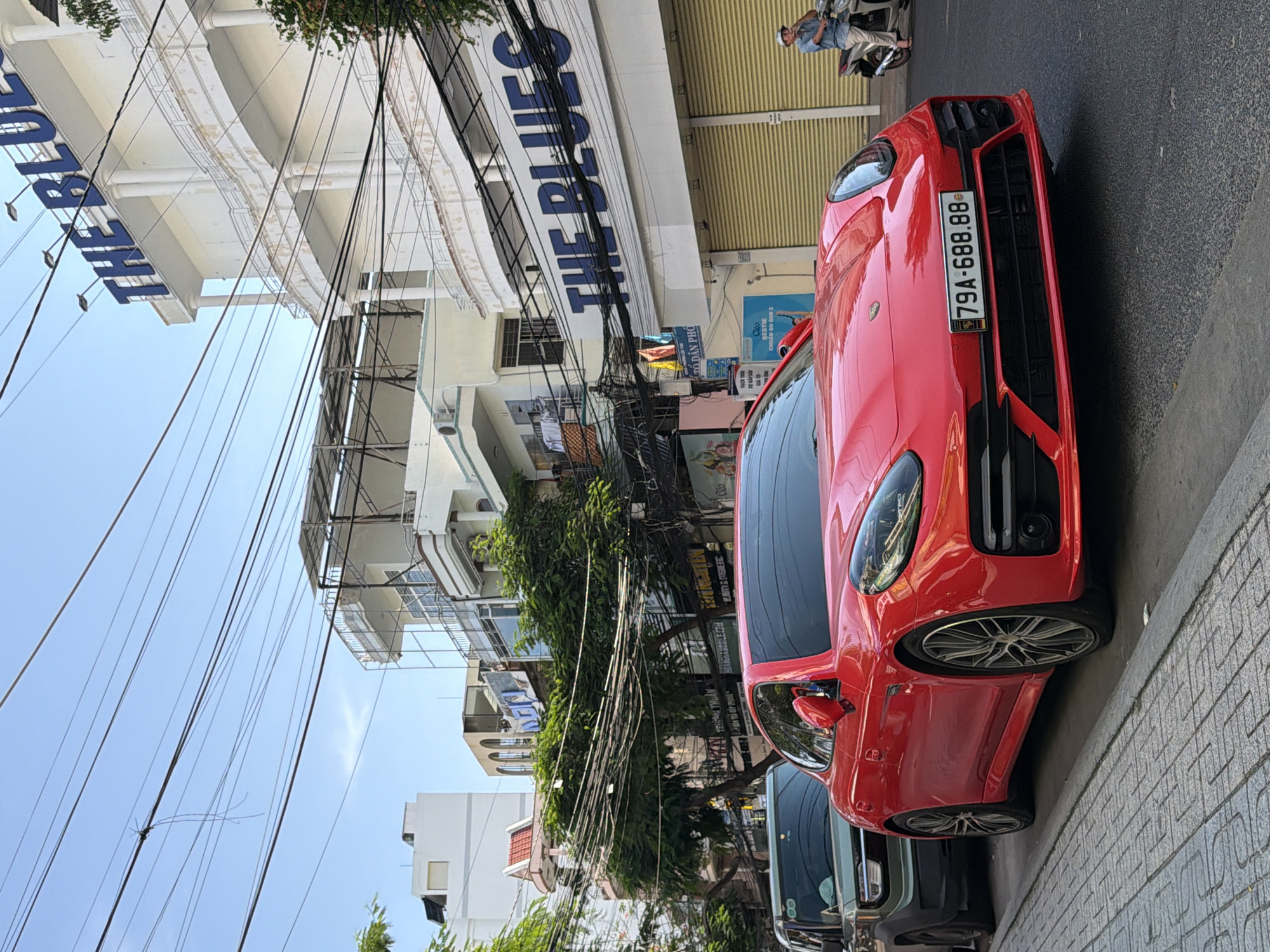 A red Porsche Panamera is parked on the side of a street in Nha Trang, Vietnam, in front of a white building with blue lettering that reads "THE BLUES". A gray SUV is parked behind the Porsche. Overhead, a tangle of electrical wires spans the sky. In the background, other buildings and trees are visible. A person on a scooter is seen in the distance to the right. The street is paved with light-colored bricks.