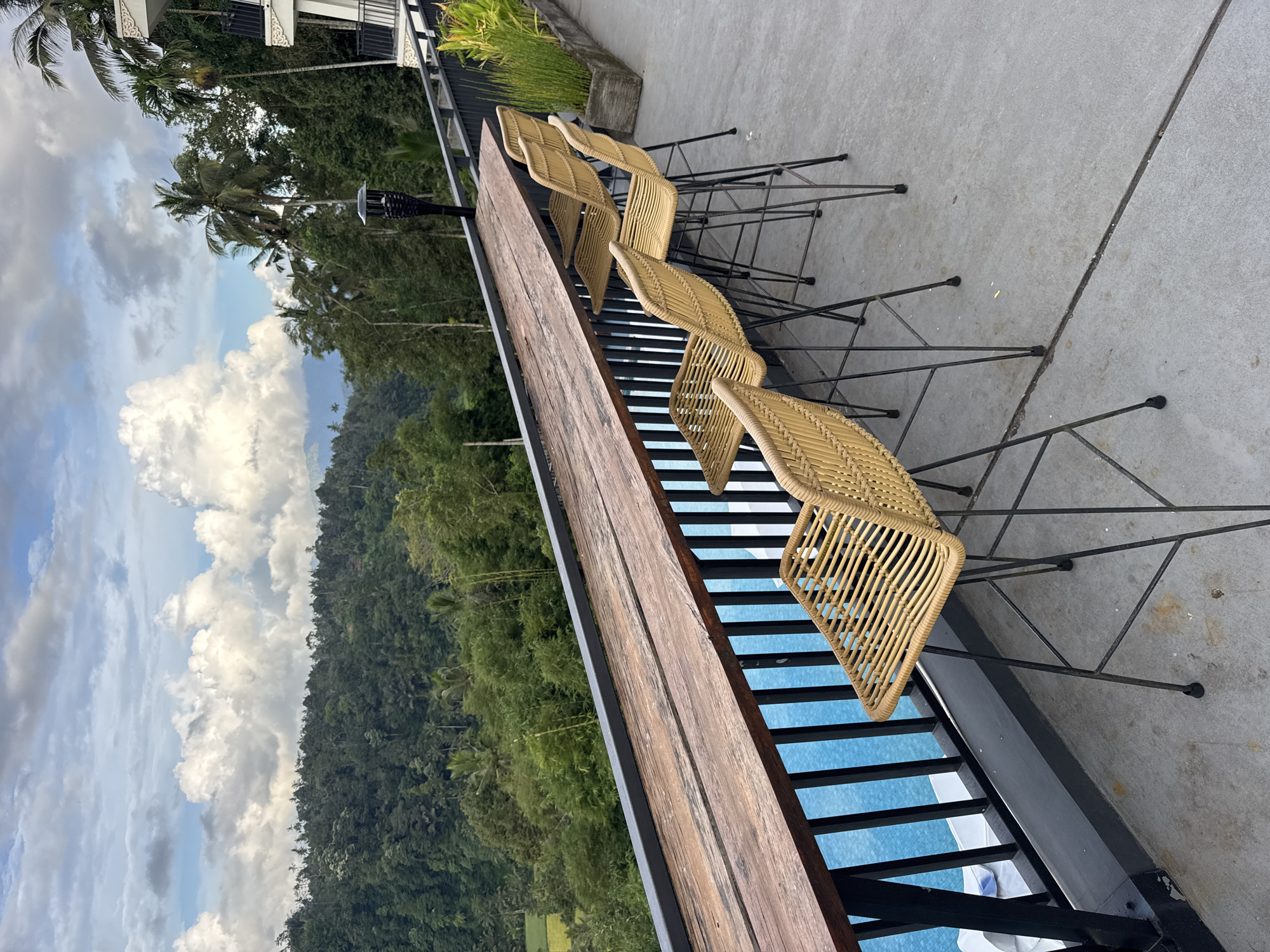 The image depicts a long, wooden table with several rattan chairs aligned beside it. The table and chairs are positioned on a concrete patio next to a railing. Beyond the railing, there is a lush, green forest and a cloudy sky. The image was taken in Rendang, Indonesia.