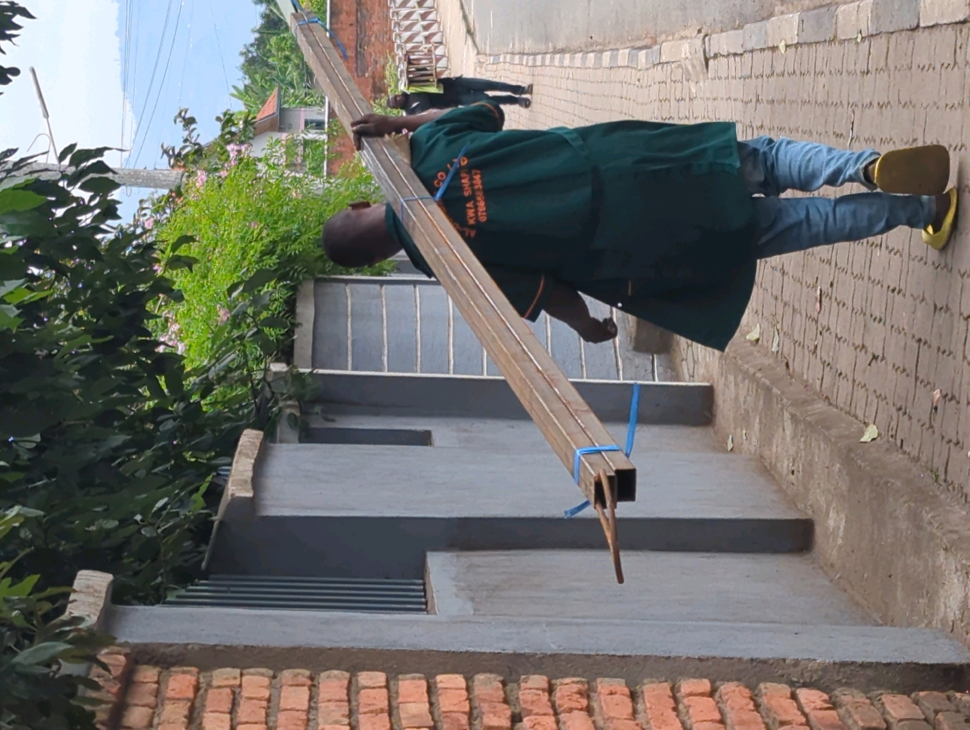 A dark-skinned man, wearing a green tunic over blue jeans and yellow slip-on shoes, is carrying a long metal beam at a diagonal across his body. The beam is secured with blue straps. He is walking on a brick-paved sidewalk in front of a gray concrete structure. In the background, there are green trees and buildings, including one with a red roof. Another person in a dark uniform is walking in the distance on the same sidewalk. The scene appears to be in a residential or commercial area in Musanze District, Rwanda. The lighting suggests it is daytime.