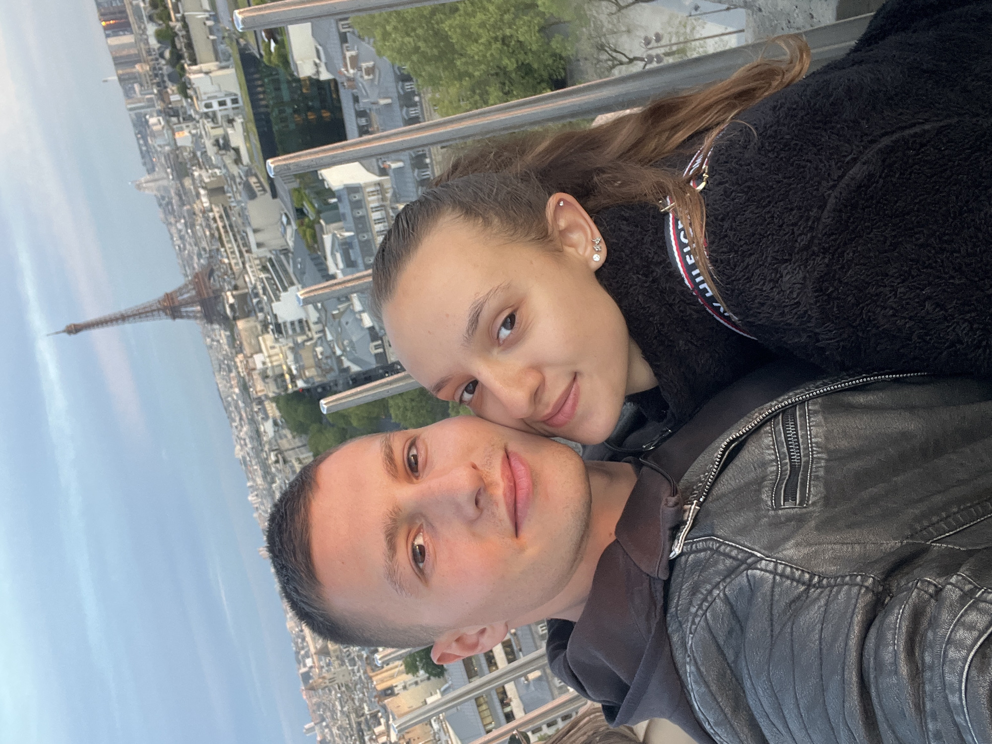 The image, originally rotated 90 degrees counter-clockwise, captures a young couple in a close-up selfie from an elevated vantage point overlooking Paris, France.
The woman, with long brown hair, is seen leaning affectionately against the man, both smiling softly towards the camera. She wears a black fuzzy jacket, with a partial view of a "TOMMY HILFIGER" logo patch on her right shoulder, featuring red, white, and blue stripes. Her ear has multiple piercings, including a small star-shaped earring. The man has short dark hair and is wearing a dark, possibly leather, jacket over a dark shirt.
Behind the couple, the distinctive silhouette of the Eiffel Tower is prominently visible against a soft, light blue sky, suggesting either late afternoon or early evening. The Parisian cityscape with its characteristic buildings and rooftops extends into the distance. Metal railings are visible in the foreground, indicating they are on a balcony or observation deck. The overall mood conveyed is one of happiness and affection, set against an iconic Parisian backdrop.