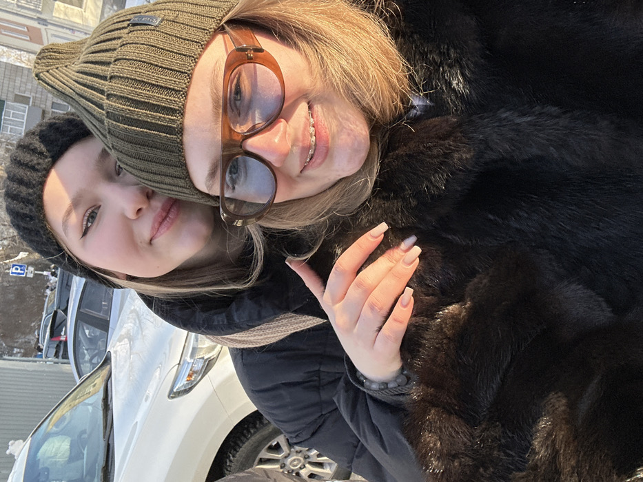 A bright daylight outdoor photograph depicts two women posing in a close embrace. The woman in the foreground on the right has blonde hair, wears an olive green ribbed beanie, large brown-framed sunglasses with light blue lenses, and a dark brown fur coat. She is smiling, revealing dental braces on her upper teeth. The woman behind her, on the left, has lighter hair, wears a black ribbed beanie, a black puffer jacket, and a light beige scarf. She is also smiling softly. Her right arm is around the woman in front, with her hand resting on the fur coat. Her nails are manicured in a light, natural color, and a dark beaded bracelet is visible on her wrist. The background features a partially visible white SUV, other parked vehicles, and snow-covered ground, indicating a cold weather environment. A blue parking sign with a white 'P' is visible in the upper background. Further in the distance, snow-covered terrain with bare trees and faint building structures are present. The scene occurs in the city of Unknown, Unknown.