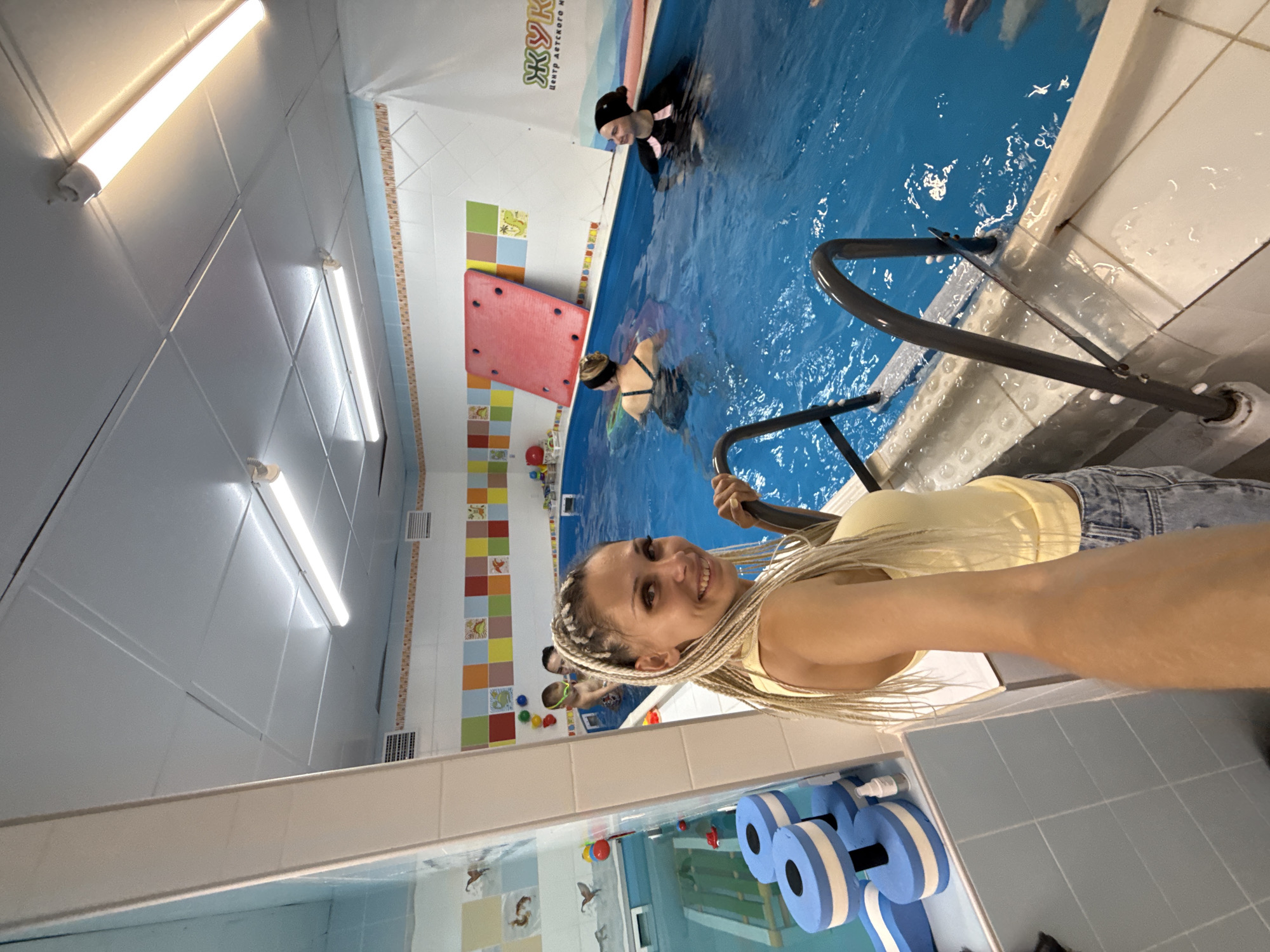 An indoor swimming pool facility. In the foreground, a woman with blonde braided hair smiles and poses for a selfie, holding onto a pool ladder. She is wearing a yellow tank top and denim shorts. In the background, several people are in the blue water. One person, wearing goggles, is being held by an adult. Another person is in a black swimsuit. The pool area is decorated with colorful tiles and aquatic-themed decals. Pool equipment, including blue and white flotation devices resembling dumbbells, is visible on the left side of the image. The ceiling is white with fluorescent lighting. A red mat is propped against the wall. A sign with Cyrillic text is visible on the right side of the pool.