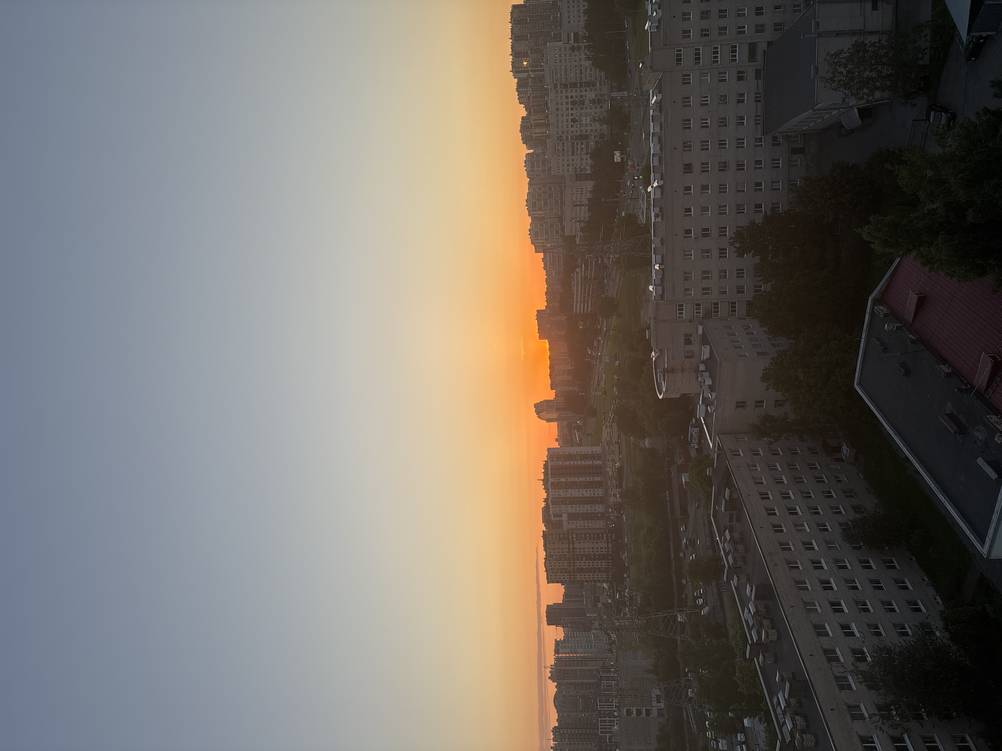 The image depicts an urban landscape at sunset or sunrise, over a "City of Unknown, Russia." The sky dominates the upper half of the frame, exhibiting a clear gradient from a pale, desaturated blue at the zenith, transitioning through light yellow, to a vibrant orange along the horizon.
Across the horizon, a dense cityscape is visible, comprising numerous multi-story residential and commercial buildings. These structures are largely silhouetted against the bright orange sky, with some closer buildings revealing details such as window patterns and facade textures.
In the midground, additional buildings, roads, and green spaces are present. Prominent power transmission towers are visible on the left side, characterized by their metallic lattice structures. Roads containing parked and moving vehicles are discernible. A large, light-colored cylindrical structure, possibly a water tower or industrial tank, stands on the right mid-ground.
The foreground features several multi-story buildings, appearing darker due to the backlighting. These buildings are predominantly grey, rectangular, and feature numerous uniform windows arranged in rows. Their flat roofs are visible, equipped with multiple ventilation units, chimneys, or HVAC systems. Clusters of dark trees are interspersed between the buildings and along presumed street lines. A portion of a dark red-tiled roof is visible in the lower-right corner of the image.
No human figures or specific human actions are clearly discernible within the frame. The overall lighting suggests a low sun angle, characteristic of dawn or dusk.