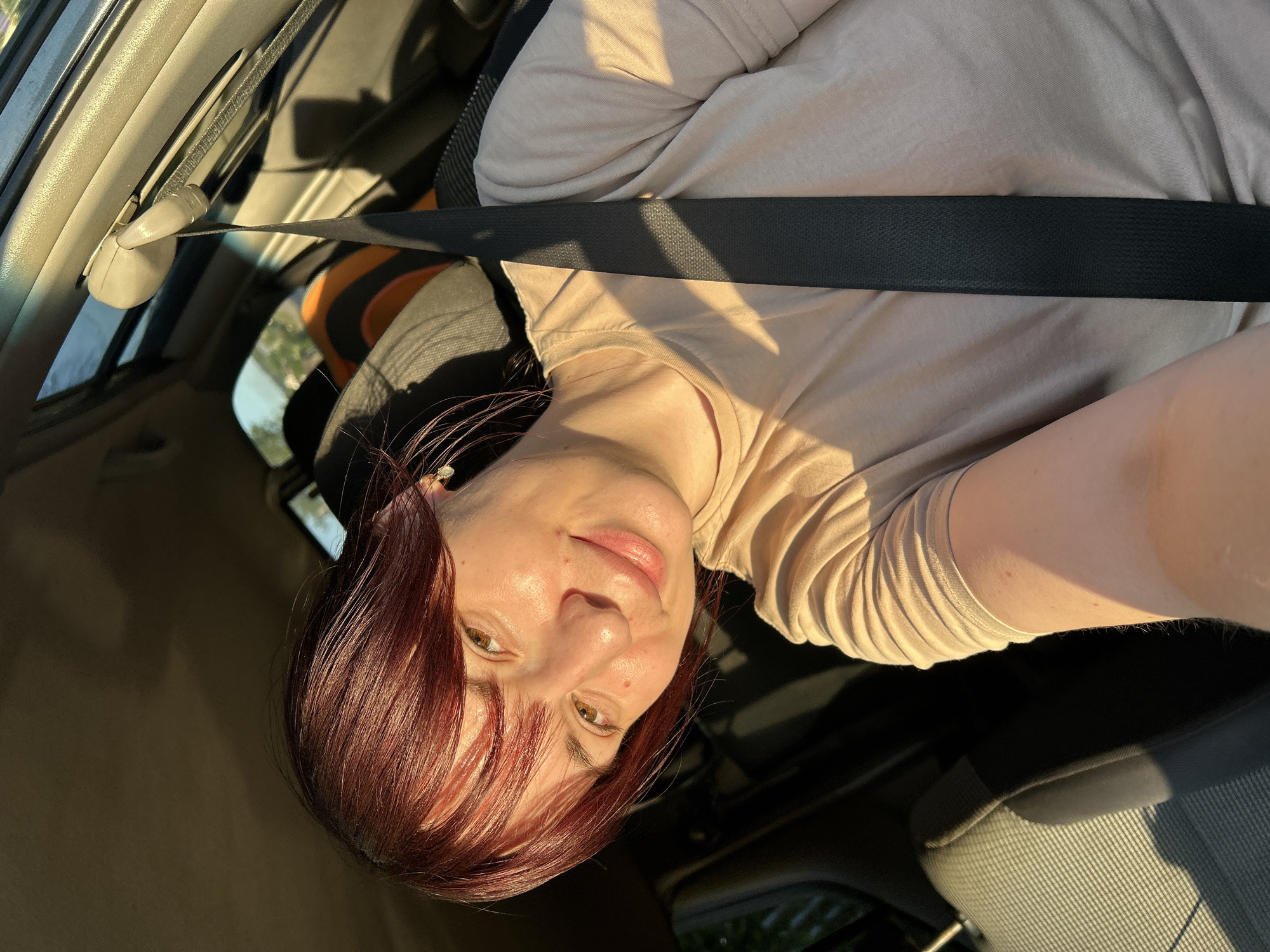 A woman with reddish-brown hair and brown eyes is taking a selfie in a car. She is wearing a light beige t-shirt and a seatbelt. The sun is shining, casting a warm glow on her face and the interior of the car. The background shows car seats and a window with a glimpse of greenery outside. The image appears to be taken during daylight. The location is not specified in the image, and therefore cannot be determined.