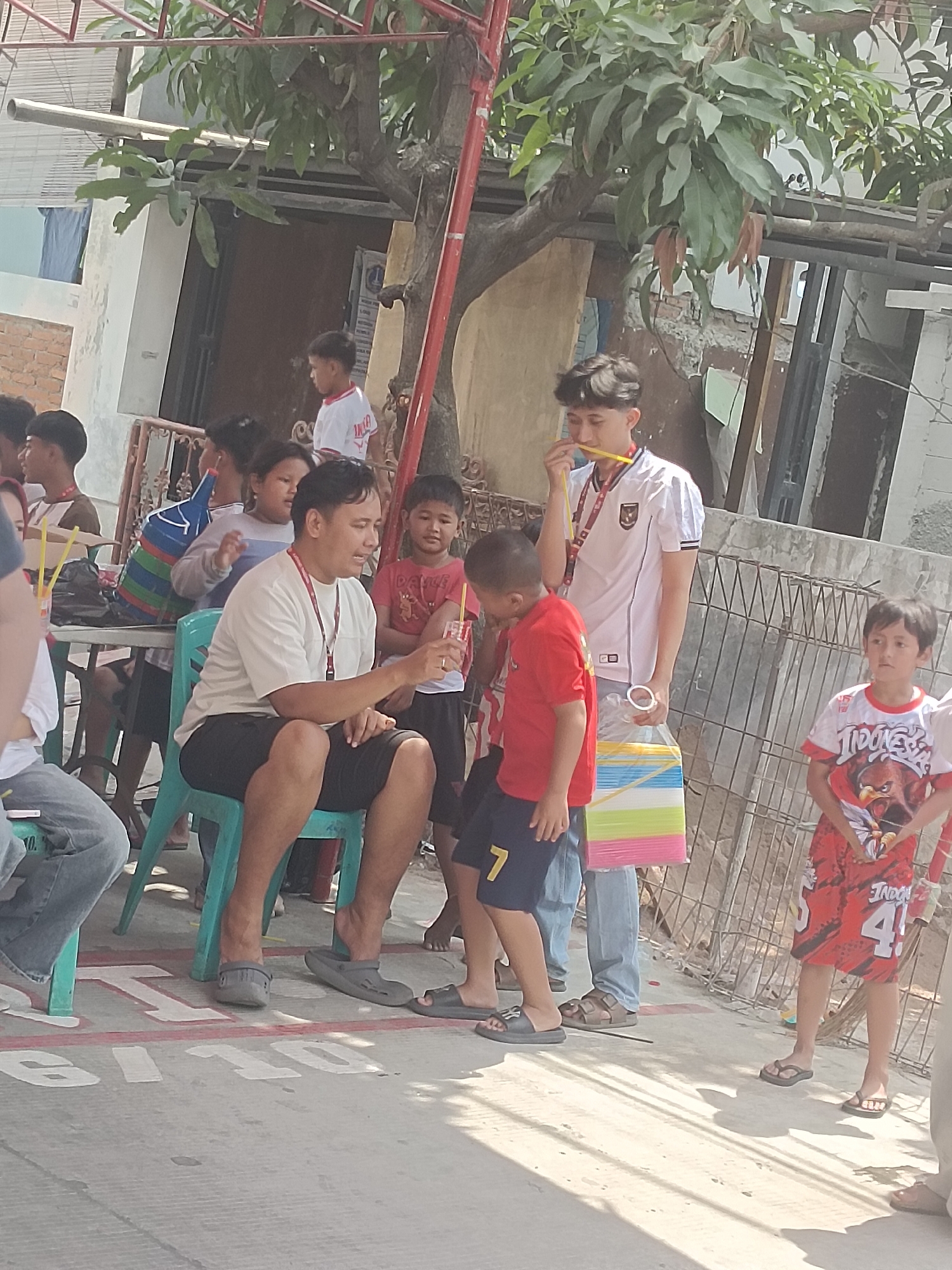 The image depicts an outdoor scene in a densely populated urban area in North Jakarta, Indonesia, likely a residential street or alley. Several people, primarily children and young adults, are present. In the foreground, a man in a white t-shirt and black shorts is seated on a teal plastic chair, holding a clear cup with a straw and appearing to be speaking. He is wearing a lanyard with a white card. To his right, a young boy in a red t-shirt with dark shorts watches him. Behind them, a young man in a white soccer jersey is standing and holding a yellow straw to his mouth, with a multi-colored bag of what appear to be straws or similar items in his hand. To the left, another child in a red and white striped shirt and dark shorts is visible. The ground is paved with concrete, marked with white lines and numbers, possibly indicating a game or activity area. Buildings with brick and plaster walls are visible in the background, along with trees and overhead structures. The overall atmosphere appears to be casual and communal, suggesting a local event or gathering.