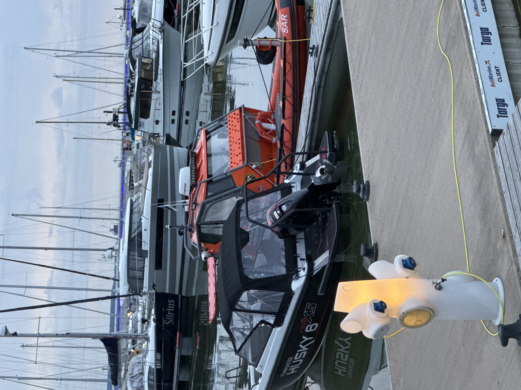This is a medium-shot, exterior, eye-level photograph taken at a harbor in Tallinn, Estonia. The scene is dominated by various boats docked in the water and on the quay. In the foreground, a white, illuminated bollard with multiple spouts and a faucet is visible, connected to a yellow hose. To the left of the bollard, a boat named "FINNMASTER HUSKY 6 XS" with a black canopy and an outboard motor is moored. Behind it, a larger boat labeled "Siirius" is docked. Further back, several other sailing boats and motorboats are visible, including one with "SAR" written on its side. The sky is overcast. The overall atmosphere is calm and industrial, typical of a harbor.