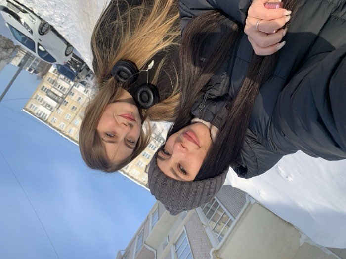 The image displays two young adult females posing for a photograph outdoors in a snow-covered urban setting in Chaikovsky, Russia, beneath a clear blue sky during daylight hours. The ground is extensively covered in snow, reflecting bright sunlight.
The female on the left has long, dark hair, wears a gray knit beanie and a black puffer jacket over a white turtleneck. She has manicured light-colored fingernails, a ring on her left ring finger, and holds a small pink object. She smiles directly at the camera.
The female on the right has long, light-brown hair, wears a dark top, and has black over-ear headphones draped around her neck. A small labret piercing is visible below her lower lip. She exhibits a subtle smile, also directed towards the camera.
In the background, multi-story residential buildings, consistent with apartment blocks, are visible. Several vehicles, including a white sedan and a dark sedan, are parked on the snow-laden ground or street to the right of the subjects. The image is captured from a low-angle perspective.