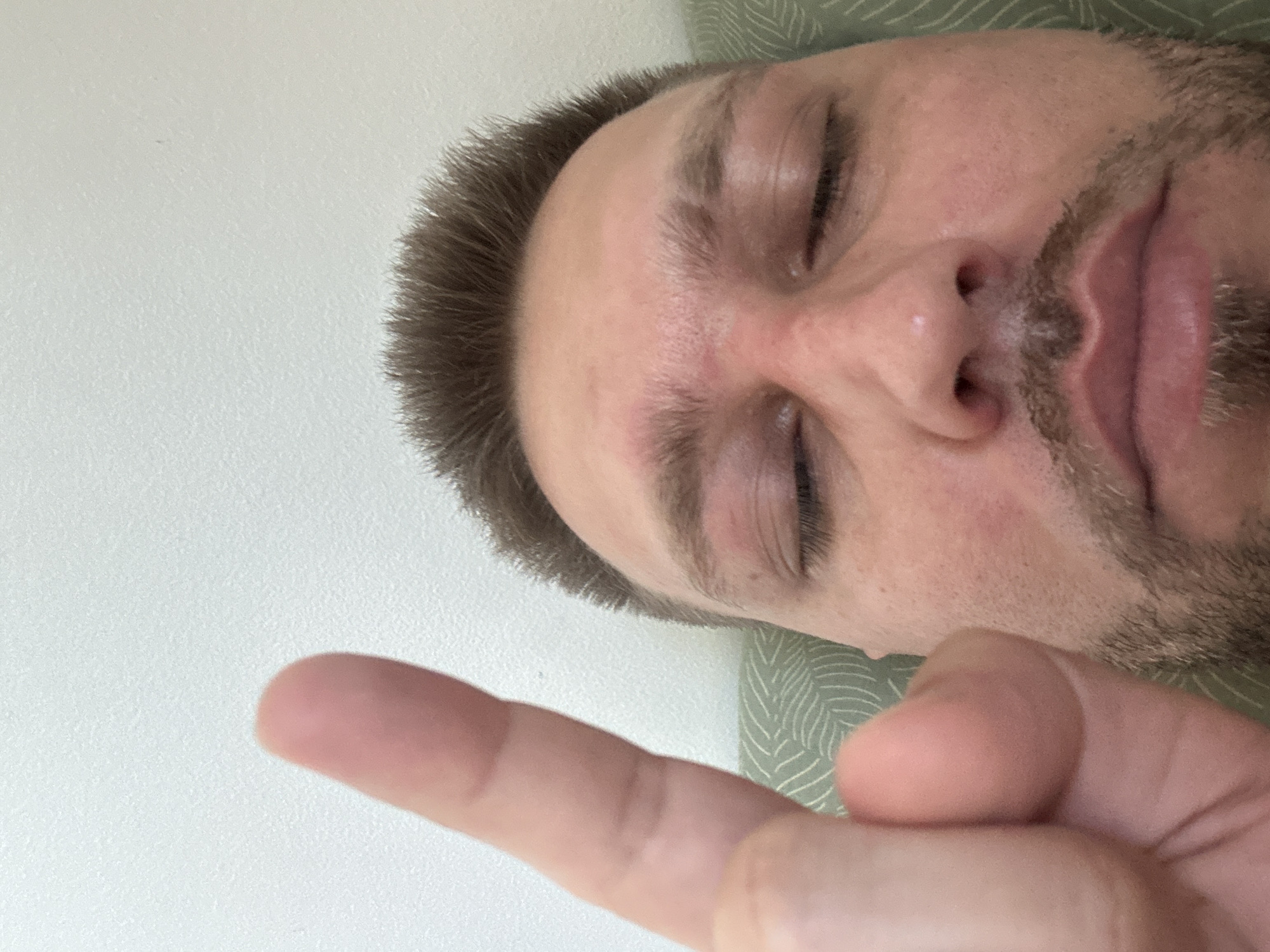 A close-up, low-angle, selfie-style shot shows a person's face with their eyes closed and their index finger raised towards the camera. The person has short, brown hair and a light beard. Their skin appears slightly red around the nose and eyes. The background consists of a textured, light-colored wall. The person is lying on a green, patterned cushion. The scene does not contain any identifiable landmarks or elements that would allow for determination of its location within Zvezdara Urban Municipality, Serbia.