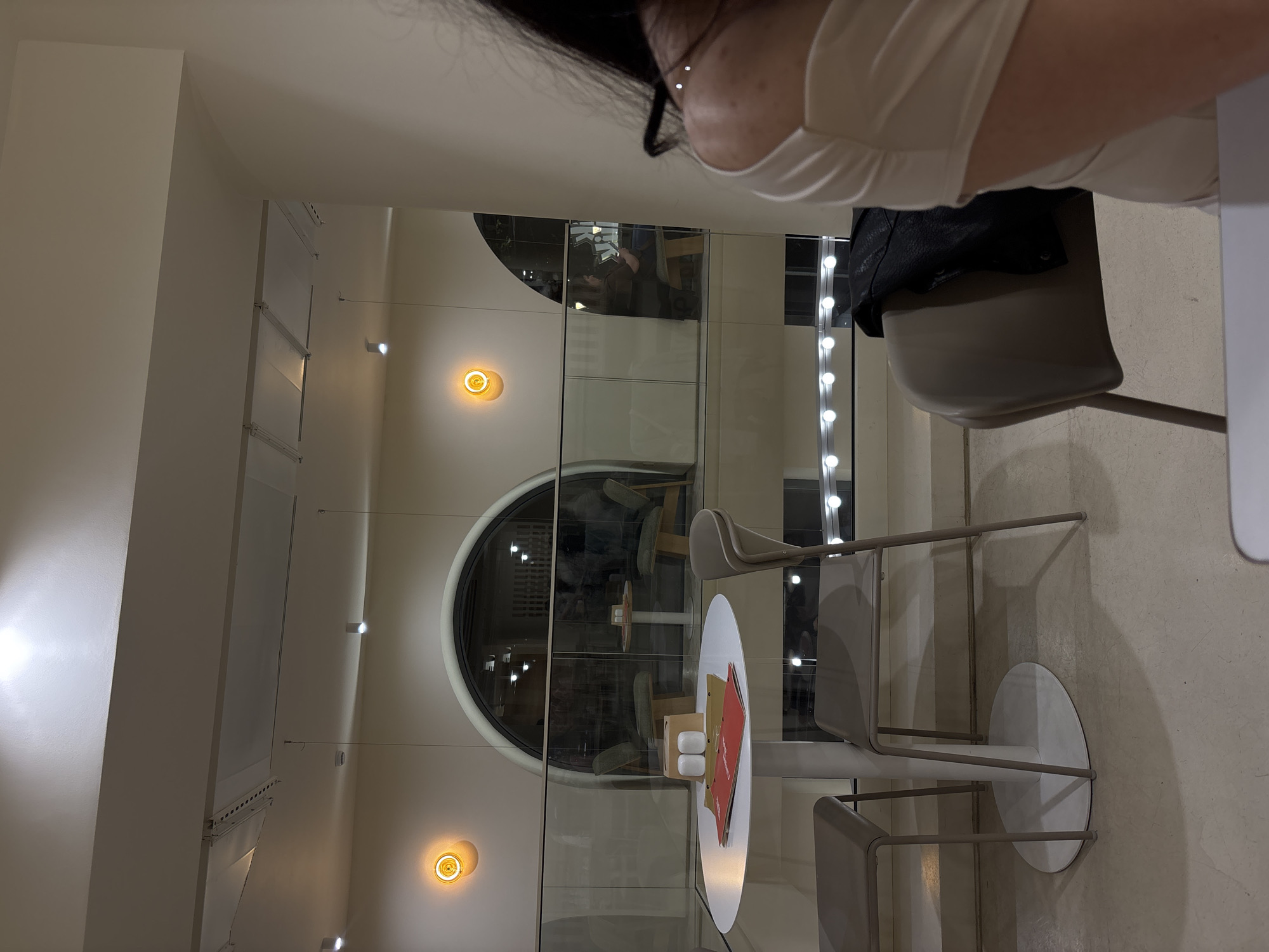 The image depicts an interior space, likely a cafe or restaurant, in Yerevan, Armenia. A masked individual is seated at a circular white table with two gray chairs. The table contains a menu and condiments. In the background, a large arched window or mirror reflects the interior, including other tables and seating. Wall-mounted lights provide illumination. The overall color scheme is muted, dominated by whites, grays, and beige tones.