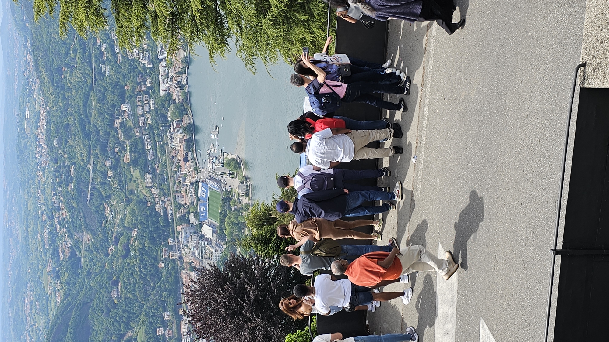 A group of approximately twelve people stand along the edge of a paved overlook at Brunate, Italy, gazing at a scenic view. The vista below includes a body of water, a city with densely packed buildings among lush greenery, and a stadium with the numbers "1907" painted on its side. The individuals are diverse in appearance and clothing, and some are taking pictures with their phones. The lighting indicates it is daytime. The setting is an elevated vantage point. A dark tree stands in the lower left corner of the frame, and a section of black metal fence is visible on the right.