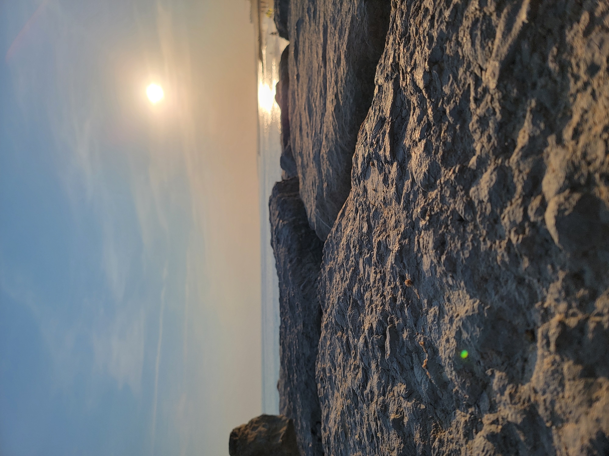 This is a low-angle, close-up photograph of rough, textured rocks in the foreground. The rocks appear to be part of a breakwater or rocky shoreline. In the background, the calm ocean stretches to the horizon, with the sun low in the sky, casting a bright, shimmering reflection on the water. The sky is a pale blue with wispy clouds. There are faint outlines of distant people on a beach or promenade to the far right. The overall impression is of a serene coastal scene at sunset or sunrise. The location appears to be Quarteira, Portugal, given the characteristic rocky coastal defenses and the shallow, calm waters often seen along this stretch of the Algarve.
