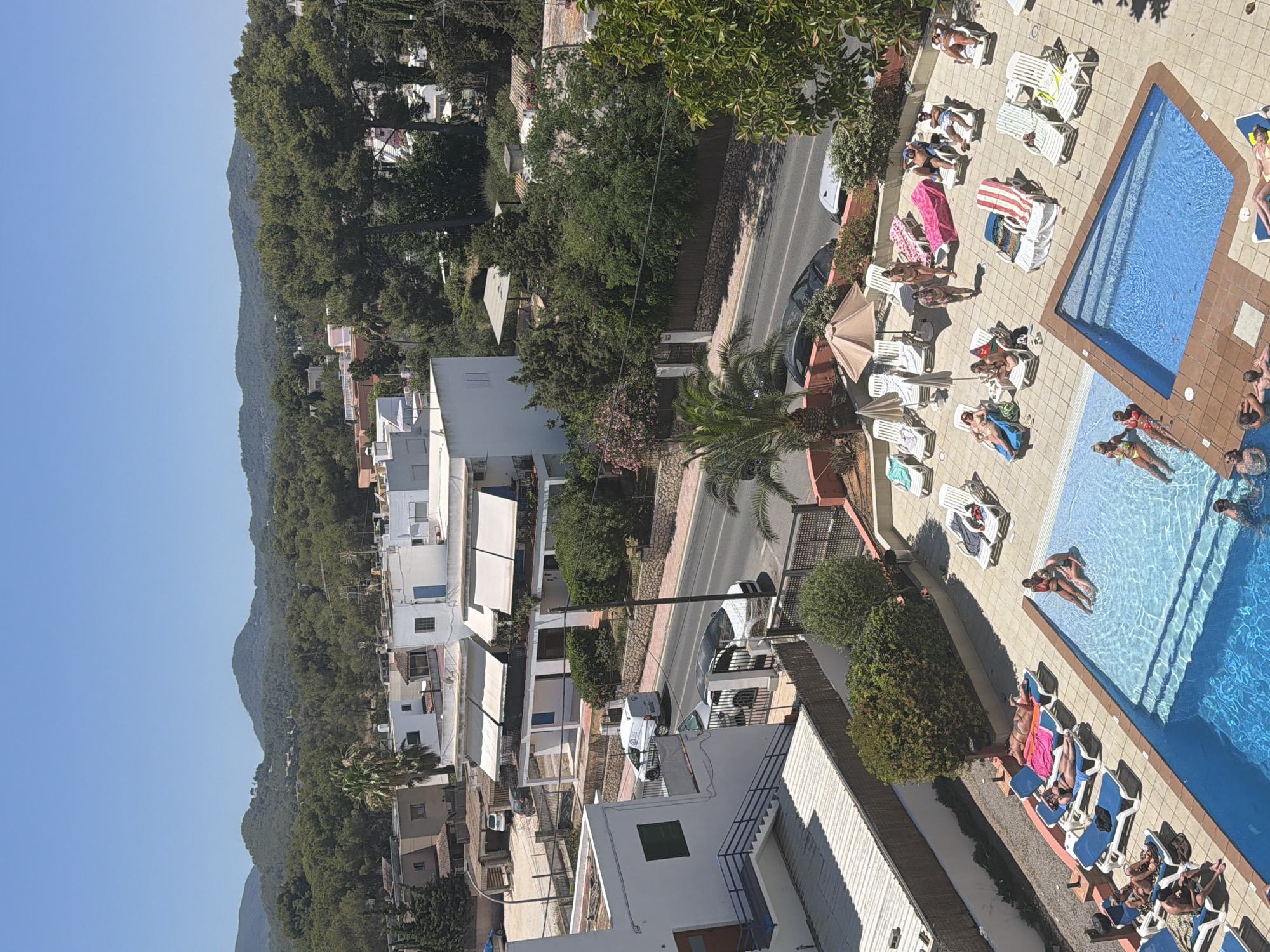 This aerial, sunlit photograph captures a scene in Sant Antoni de Portmany, Spain, on a clear, cloudless day. The image focuses on a resort-like setting with a swimming pool and surrounding lounge chairs. In the foreground, a large blue swimming pool is partially visible, with several people enjoying the water. One group of three women in swimsuits are relaxing near the edge of the pool. Further into the pool, two men are visible, one with his head above water and another possibly submerged.
Surrounding the pool is a tiled patio area with numerous white lounge chairs. Some chairs are occupied by individuals sunbathing or resting. On one chair, a person lies face down, another is seated with a towel, and a couple sits together on a lounger. Various beach towels in bright colors like pink and blue are scattered on the chairs. Several umbrellas provide shade over some of the seating areas.
Beyond the pool area, a paved road runs horizontally across the mid-ground, with a few cars and a white van visible. Buildings with white facades and balconies with awnings are situated along the road. Green trees and vegetation are abundant throughout the scene, with palm trees and taller evergreen trees framing the buildings and the distant hills. The background features rolling green hills and a clear blue sky. The overall impression is one of a vibrant summer vacation destination.