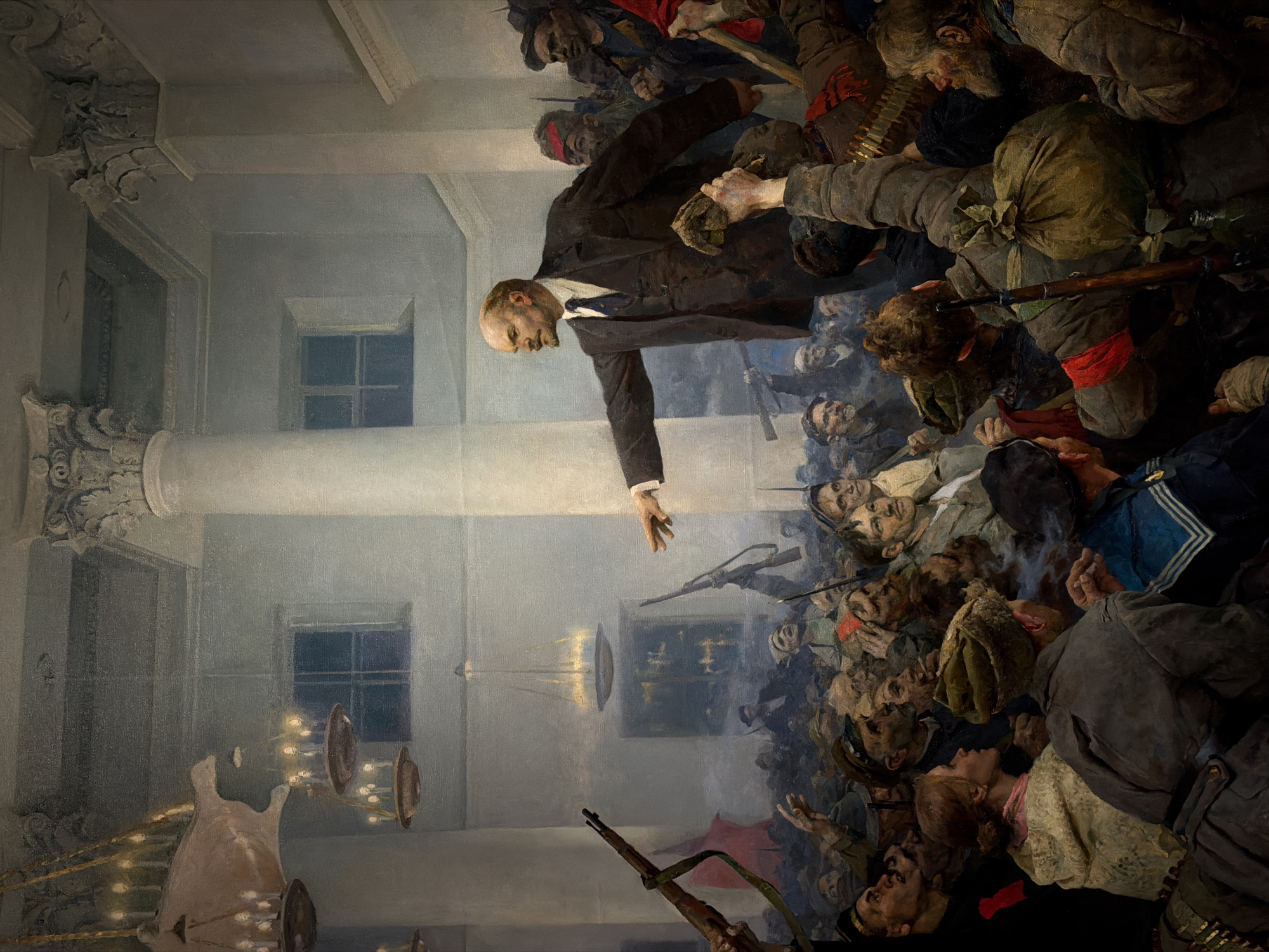 This is a painting depicting Vladimir Lenin addressing a crowd of revolutionary soldiers and civilians inside a grand hall. The hall features large windows, ornate columns, and chandeliers. Lenin, dressed in a dark suit, stands on a platform, extending his hand towards the crowd. The crowd, composed of men in military uniforms, sailors, and civilians, brandish rifles and other weapons. The scene conveys the energy and fervor of a revolutionary moment. The city is not explicitly mentioned, but the painting is generally associated with events from the Russian Revolution in Petrograd (now St. Petersburg), Russia, not Moscow.
