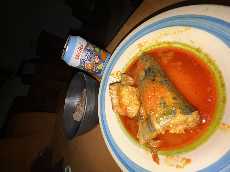 This image, captured in Egbe, Nigeria, displays an indoor dining scene. A white circular plate with blue and green striped rim contains a meal consisting of a large portion of fish and two smaller protein pieces, all coated in a vibrant reddish-orange sauce. To the left of the plate, a black bowl holds a quantity of greyish-brown, solid food. An opened, light blue and white Chivita Active Zest Mixed Fruit Drink can is positioned to the right of the bowl. All items rest on an orange-brown surface. The background is dark and out of focus, suggesting an interior space with dim ambient lighting and indistinct furniture.