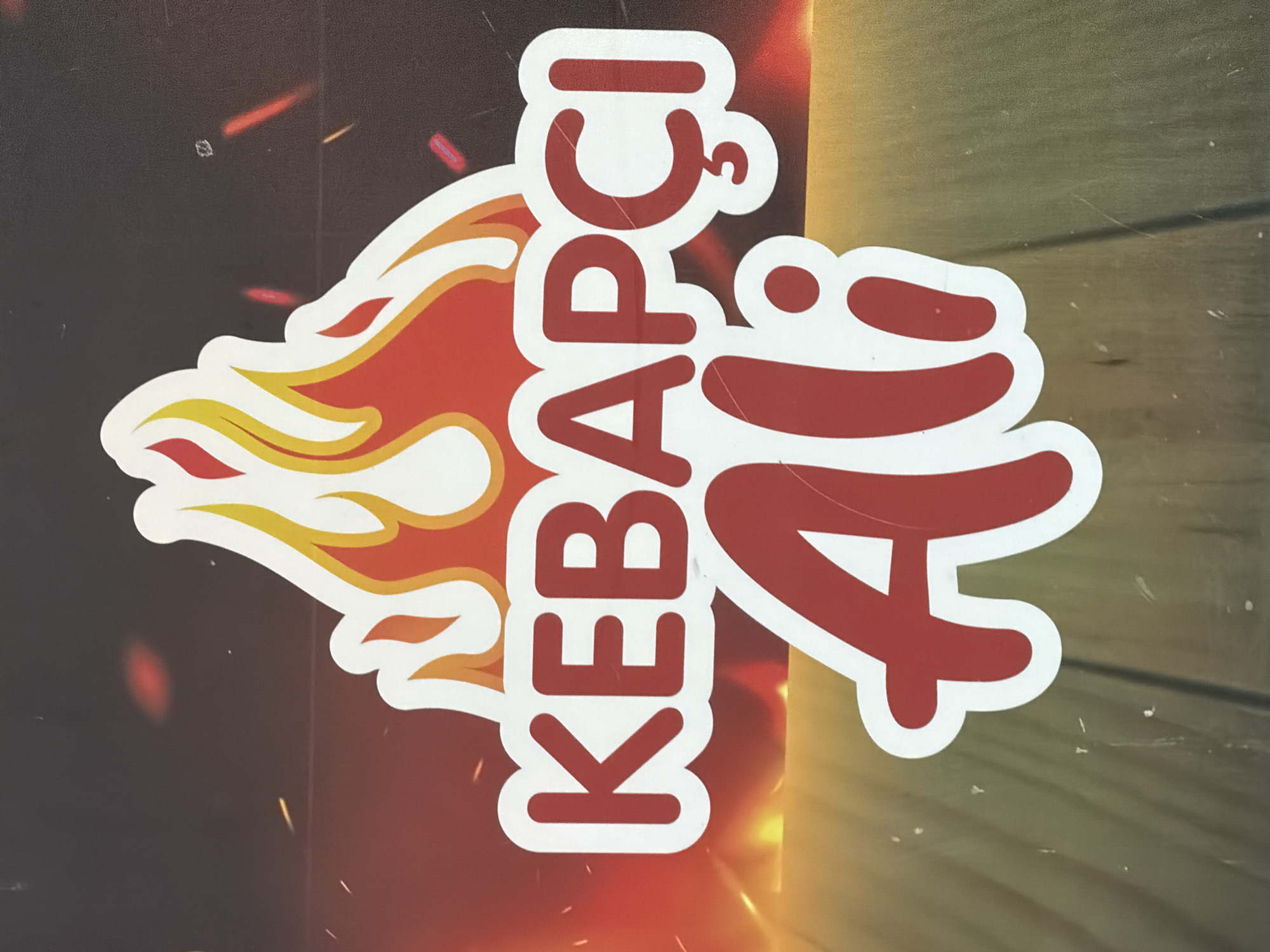 The image displays a logo with stylized flames in red and yellow. The text "KEBAPÇI" is written in large red letters with white outlines, positioned above "Ali" which is written in a larger, flowing red script with a white outline. The logo appears to be on a dark background with subtle fiery elements and what looks like a wooden texture at the bottom. The logo signifies a kebab restaurant. There is no specific location in Kocaeli, Turkey identified in the image.