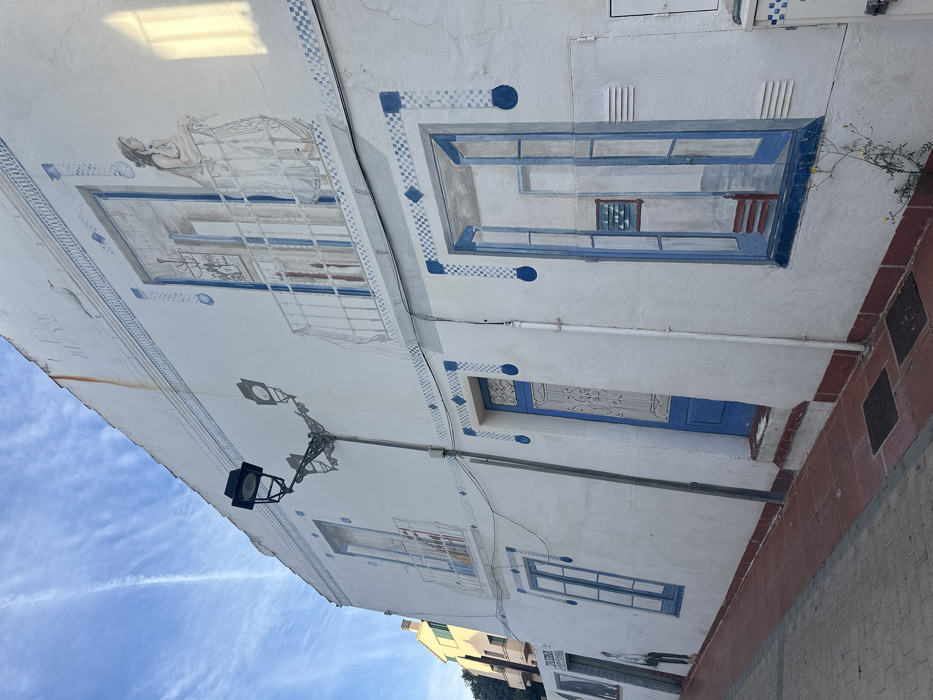 The image displays a white building facade in Calella De Palafrugell, Spain, extensively adorned with blue and white trompe l'oeil murals under a clear blue sky with faint contrails. A prominent black, ornate street lamp with two light fixtures is affixed to the upper half of the wall, left of center.
On the upper level, painted architectural features include two windows on the left and a central balcony with an open door, through which a female figure in a white dress leans on the railing, observing a small white bird on the balcony floor. Blue checkered and circular patterns border these painted elements and form horizontal decorative lines across the facade. Faint rust streaks are visible on the upper wall.
The lower level features three distinct painted sections: two blue windows on the far left, a central blue door with white ornamental detailing, and a large painted open window on the right revealing an interior scene with a red chair and a framed picture. Vertical white pipes run along the wall between these elements. The ground level presents a reddish-brown tiled sidewalk with two dark rectangular doormats. A small plant with yellow flowers grows at the base of the wall on the right. A partial view of another light-colored building is visible in the background on the far left.