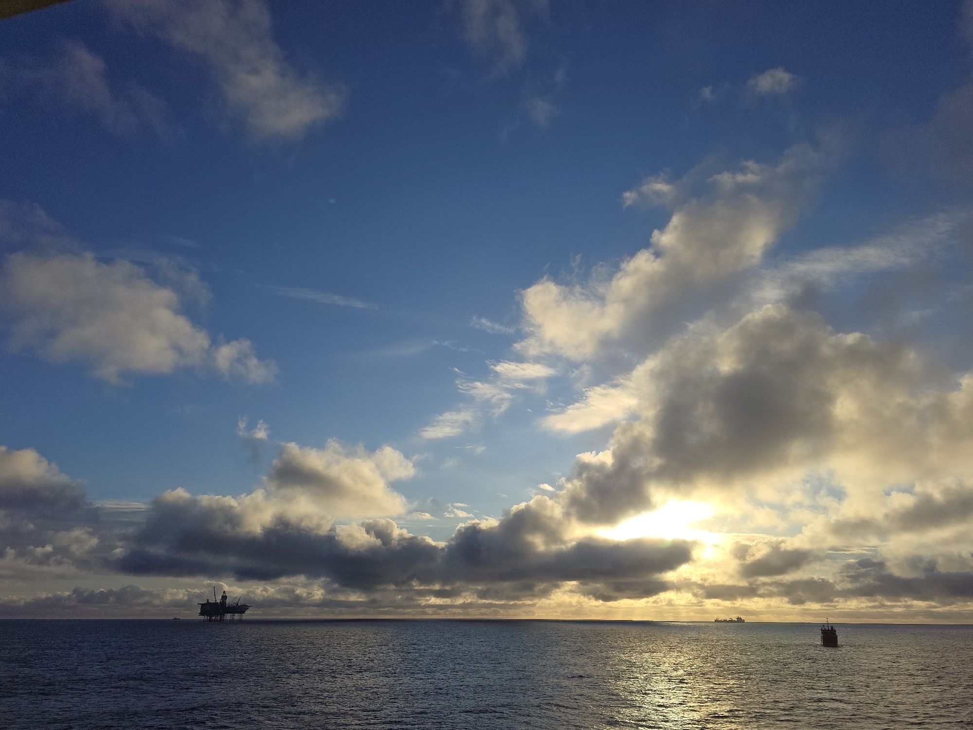 The image shows a seascape with a cloudy sky and a calm sea. An oil rig is visible on the left side of the horizon. A small boat is visible to the right of the oil rig. The sun is setting, casting a golden glow on the water. The image appears to be taken at sea near Randaberg, Norway, given the presence of the oil rig.