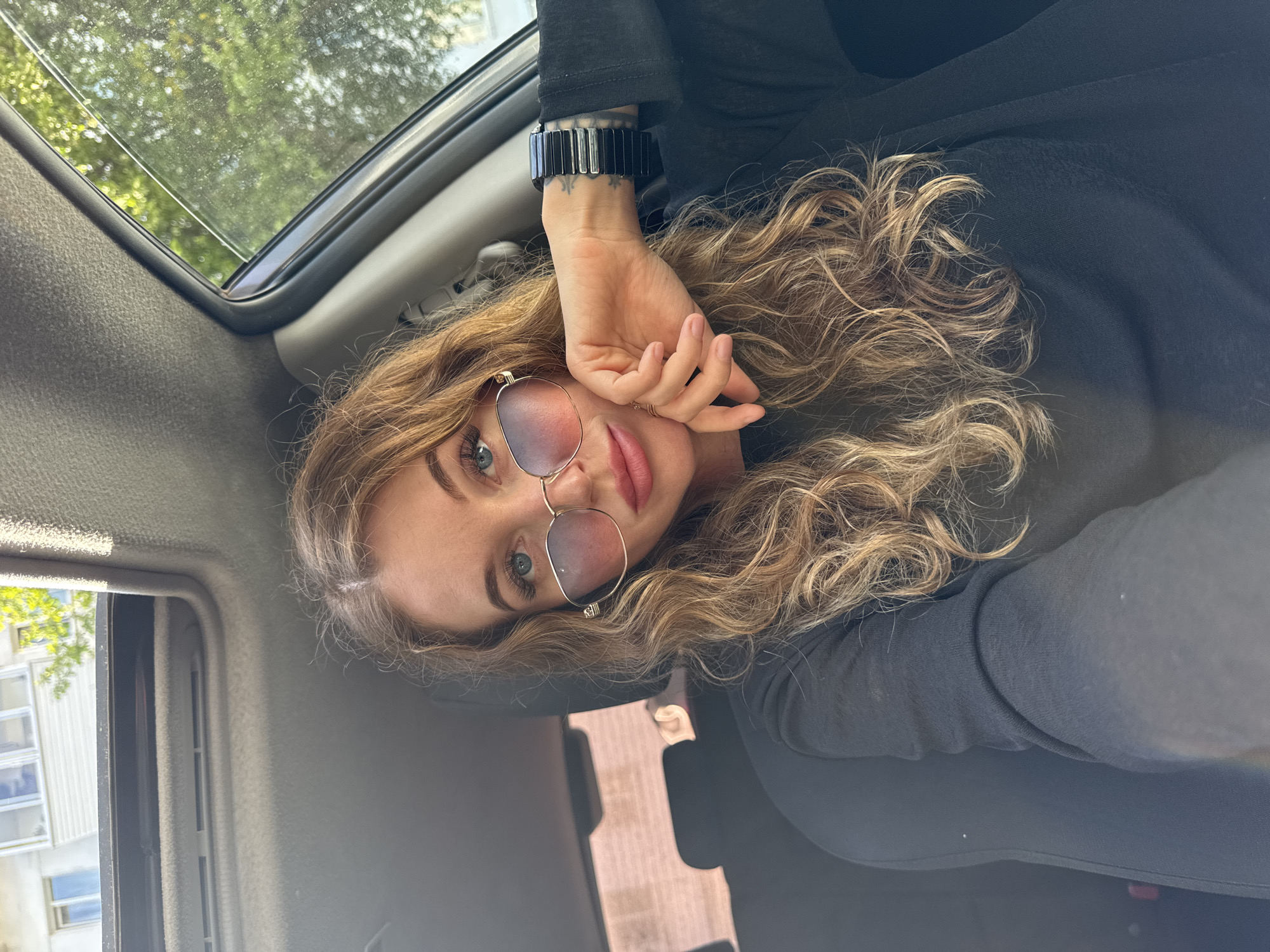 A woman with blonde curly hair, wearing sunglasses and a black top, is lying in a car, presumably with a sunroof, looking at the camera. The image is rotated counterclockwise, revealing the car's interior, including the sunroof showing trees and sky, and a side window with a glimpse of a building. The woman's left hand is resting on her face, and she is wearing a bracelet on her left wrist. The location in Chaikovsky, Russia, cannot be determined based solely on the provided image.