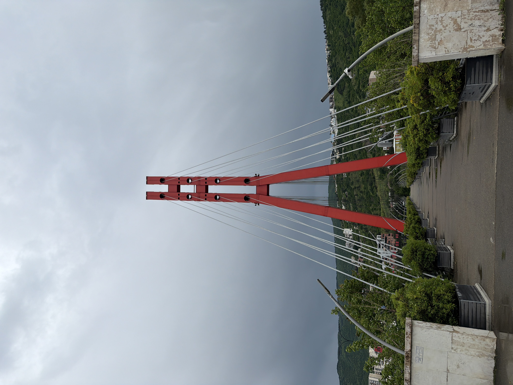 The image depicts the Bridge of Peace in Tbilisi, Georgia, on a cloudy day. The red support structure of the bridge is prominently displayed, with white cables extending from it. The bridge pathway is visible, lined with greenery in planters. The background shows the city landscape and forested hills.