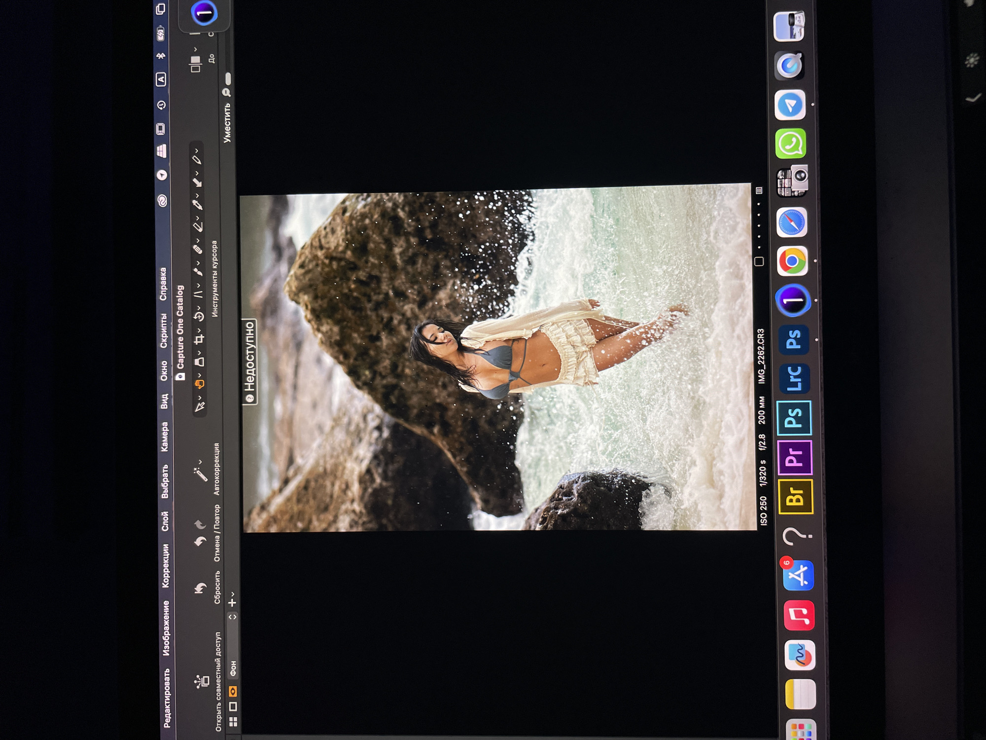 The image shows a photograph of a woman in a bikini and a light-colored shawl lying in shallow water near rocks. The photograph is displayed on a tablet screen, likely an iPad, which also shows the user interface of the "Capture One Catalog" photo editing software. The tablet screen also displays the MacOS dock with a variety of application icons.