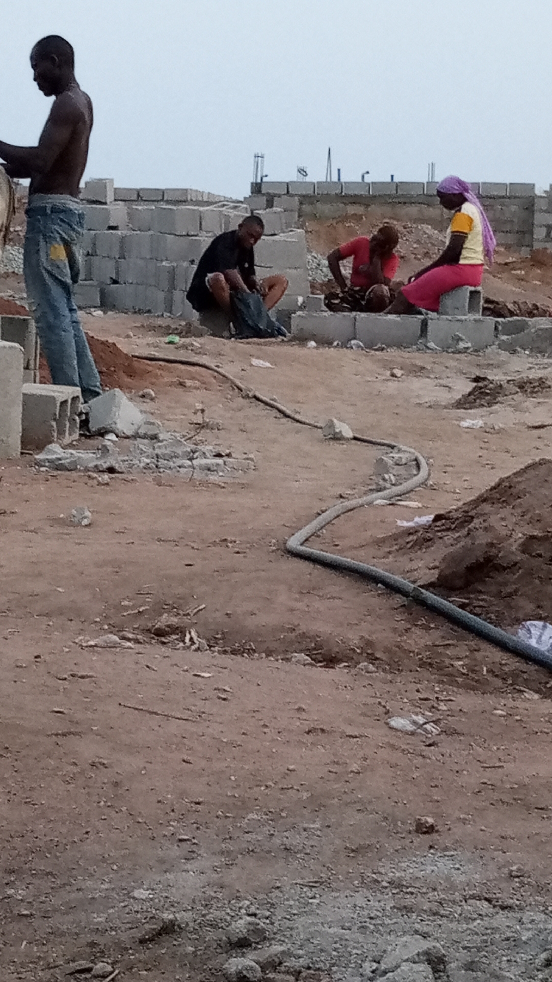 The image depicts an outdoor construction site in Adankolo, Nigeria. There are four individuals present. One man, shirtless and wearing jeans, stands on the left side of the frame. A second man in a black shirt sits amongst the cinder blocks. Two women are seated nearby. One wears a pink skirt, yellow top, and lavender head wrap, while the other wears a red top. All three seated individuals are engaged in a task, possibly related to the construction. Cinder blocks surround them. A black hose snakes across the dirt ground. The environment appears to be a building site with exposed earth and scattered debris. The sky is overcast.