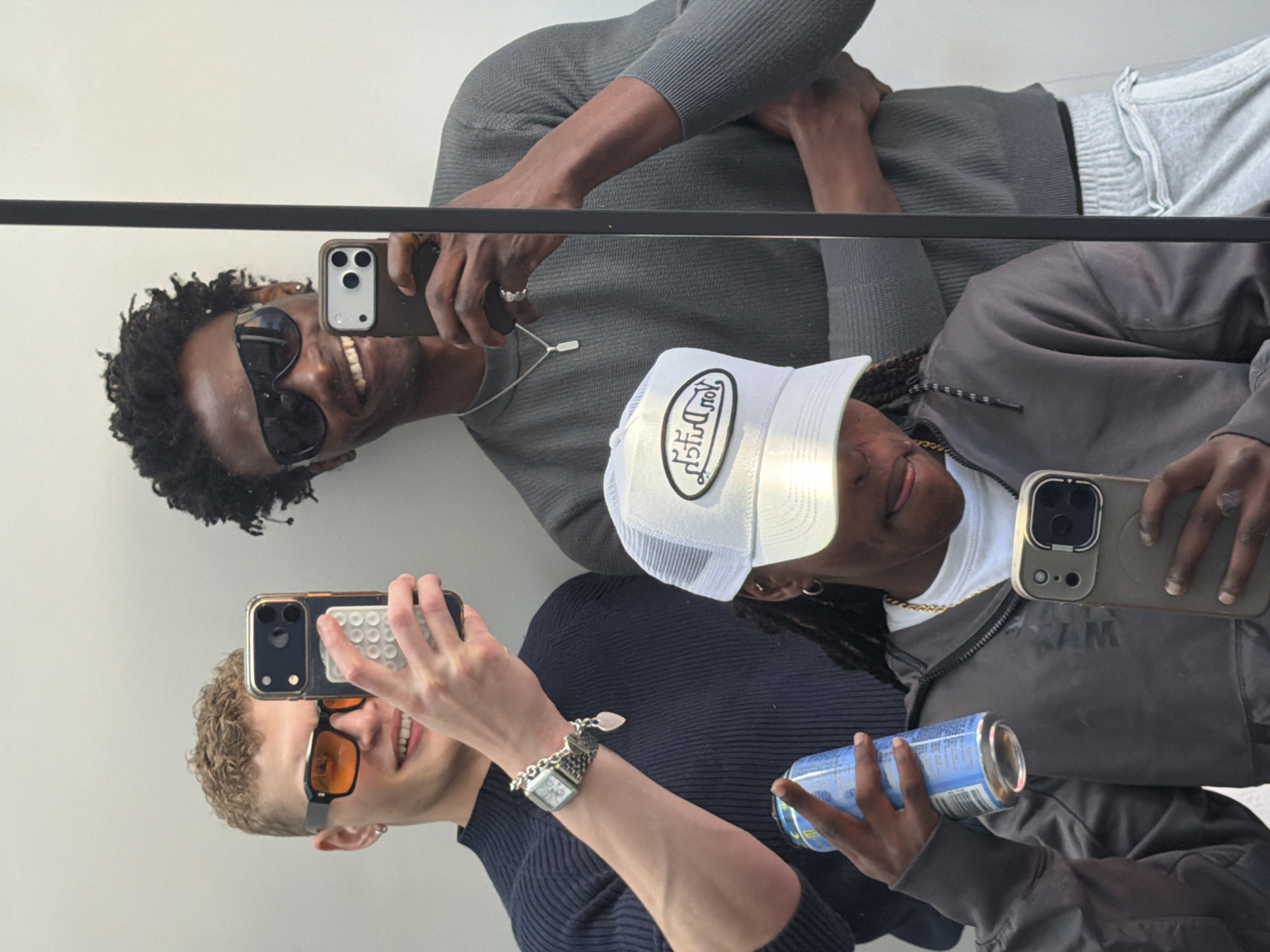 Three individuals are taking mirror selfies, presumably in London. The leftmost person is light-skinned with short curly hair, wearing orange-tinted sunglasses, a navy ribbed top, and a watch. They are holding a phone with a studded case. Adjacent to them, another person of color is partially visible, wearing a white trucker hat, a gold chain, a white t-shirt, and a gray bomber jacket; they are holding a blue can. Above them, another person of color with curly hair is wearing sunglasses, a grey top, and a silver necklace, holding a phone. The mirror has a black horizontal bar running across it. The background is a plain white wall.