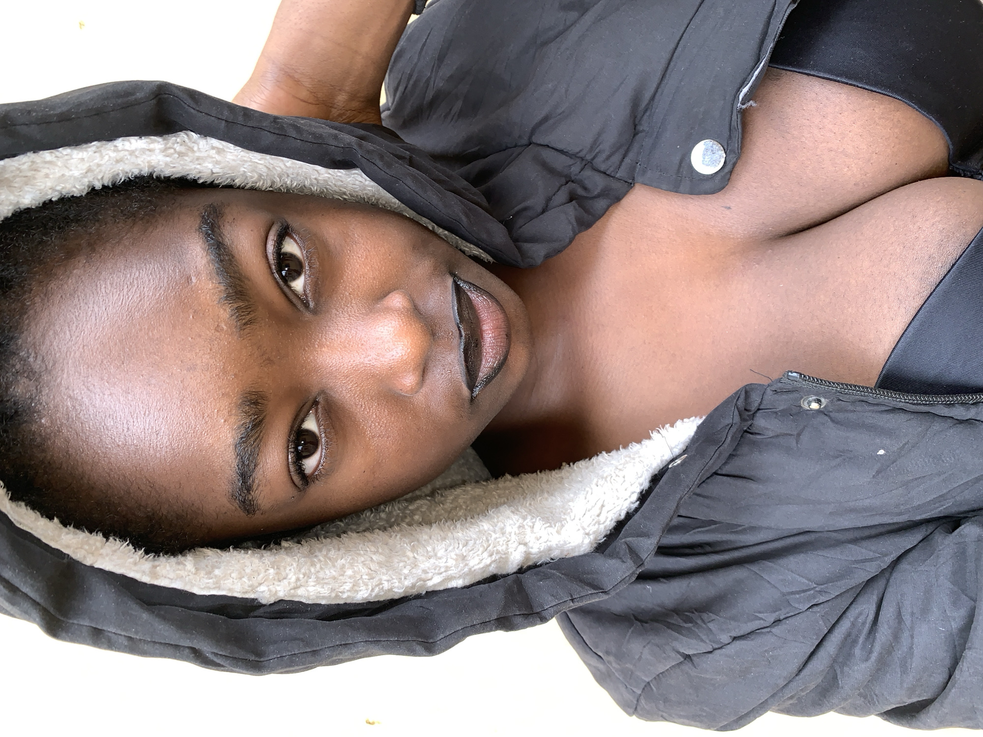 A close-up, portrait-style photograph shows a Black woman, possibly lying down, facing the camera. She wears a black jacket with a white fleece lining around the hood. Her skin is visible from the collarbone up, revealing a black top. The background appears to be a plain, light-colored surface. The location in Rafin Par, Nigeria is not discernible from the image.