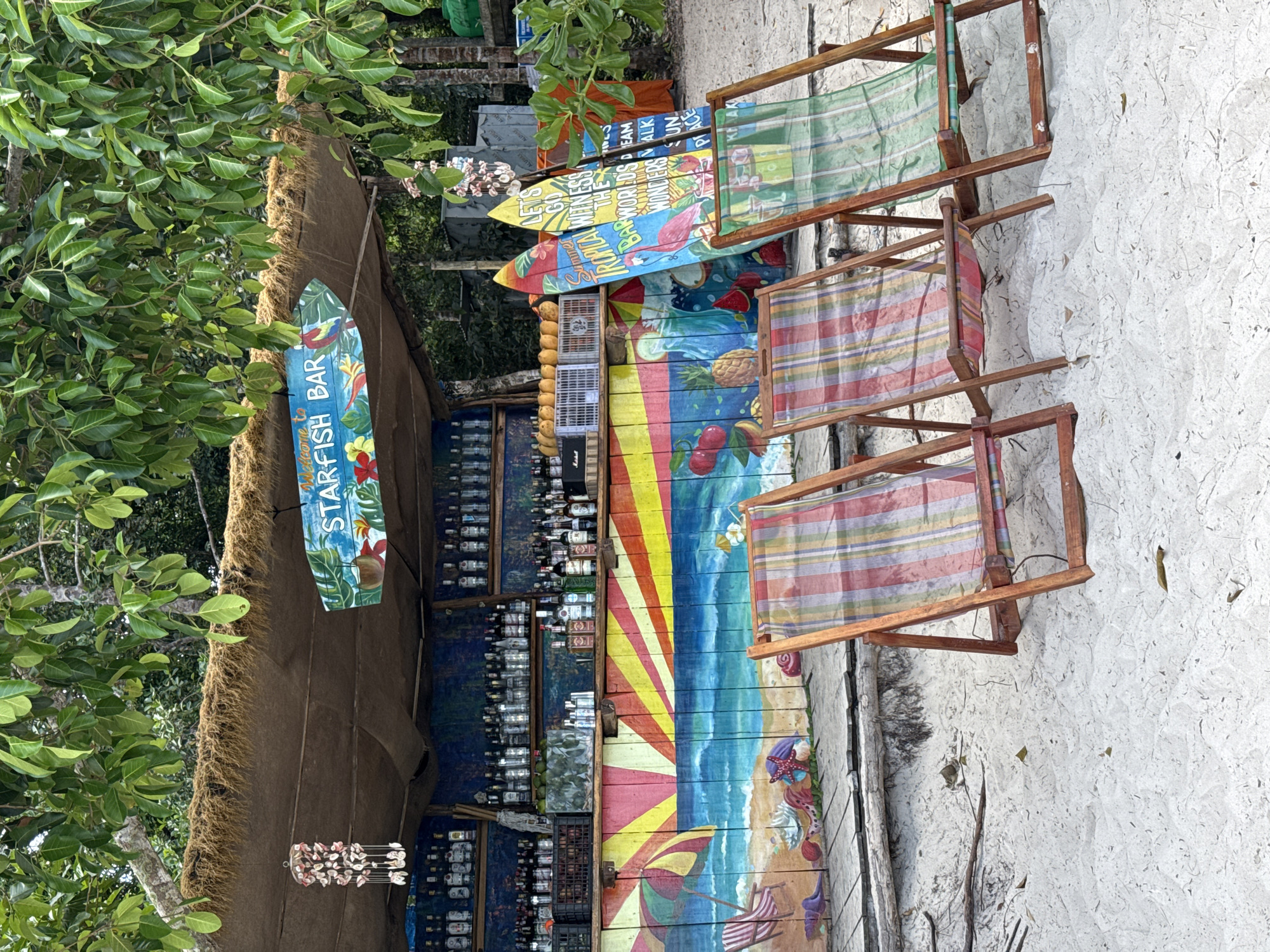 The image shows a beachfront bar in Phú Quốc, Vietnam. The bar has a thatched roof and a sign reading "Starfish Bar." Various colorful signs are displayed, one reading "LET'S GO TROPICAL WITNESS THE BARWORLDS DREAM WONDERS WALK SUN PLACE." A selection of bottled beverages lines the back wall of the bar. A mural depicting a beach scene, including a beach umbrella and a striped beach chair, is painted on the front. Three wooden-framed beach chairs are arranged in front of the bar on the sandy beach. Lush green foliage is visible to the left of the bar.