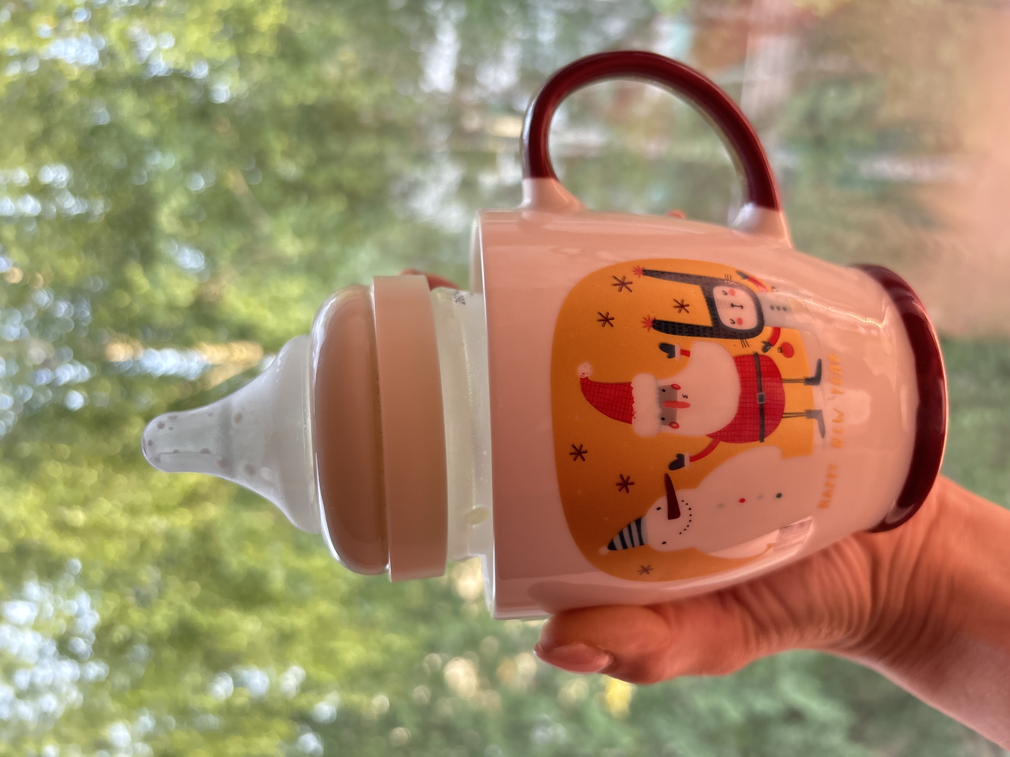 A person is holding a white mug with Christmas-themed cartoon drawings attached to a baby bottle. The mug features a brown handle and images of Santa Claus, a stick figure angel, and a snowman on a yellow background. The baby bottle is attached to the mug's rim, containing a clear liquid. The hand holding the mug appears to be pale-skinned. The background consists of a blurred outdoor scene, presumably a garden or trees seen through a window, out of focus. The photo might have been taken in the city of Chaikovsky, Russia, but the image itself provides no concrete evidence of this location.
