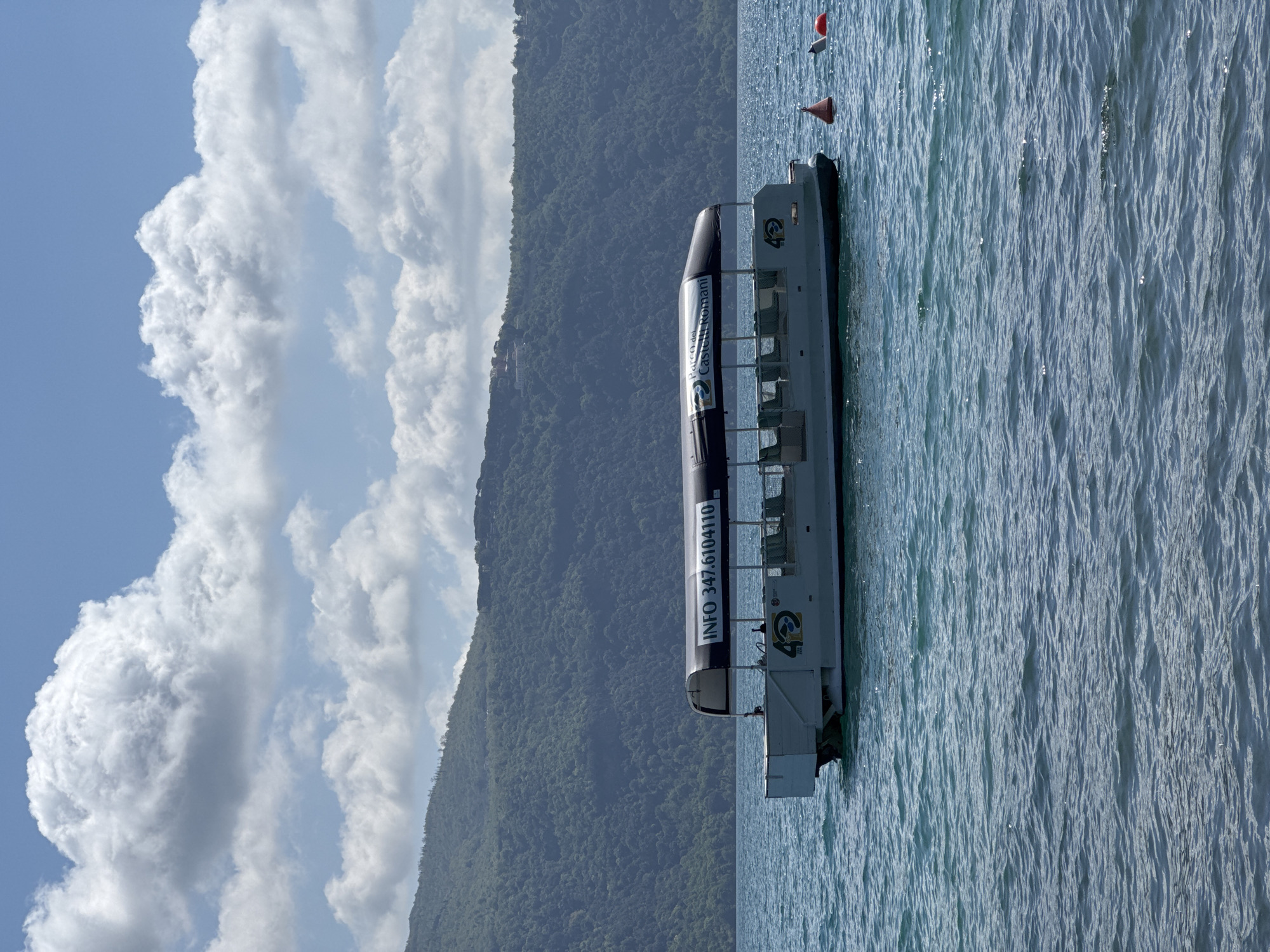 This image depicts a serene daytime scene featuring a boat on a body of water, with forested hills in the background and a partly cloudy sky above. The perspective is from the water level, looking towards the shore.
In the foreground, a white and black open-sided passenger boat floats on the calm, blue-green water. The water's surface exhibits gentle ripples and shimmering glints from the bright sunlight. Two buoys, one red conical and one orange round, are visible in the water to the right of the boat. The boat has multiple rows of open seating, suggesting it is a sightseeing or tour vessel.
Text clearly visible on the black upper section of the boat reads "Parco del Castelli Romani", identifying the regional park where it operates, and "INFO 347.6104110", likely a contact number. A logo, possibly an intertwined '42' or '4G' with "S.R.L." below it, is present on the white hull near the boat's front. No people are visible on the boat or in the immediate vicinity.
The mid-ground and background are dominated by densely forested green hills or mountains rising from the water, extending across the horizon. Above these hills, the sky is a clear light blue, dotted with large, fluffy white cumulus clouds. The bright illumination indicates it is either late morning or early afternoon. The scene conveys a sense of tranquility and natural beauty, consistent with a scenic lake environment such as Castel Gandolfo in Italy.