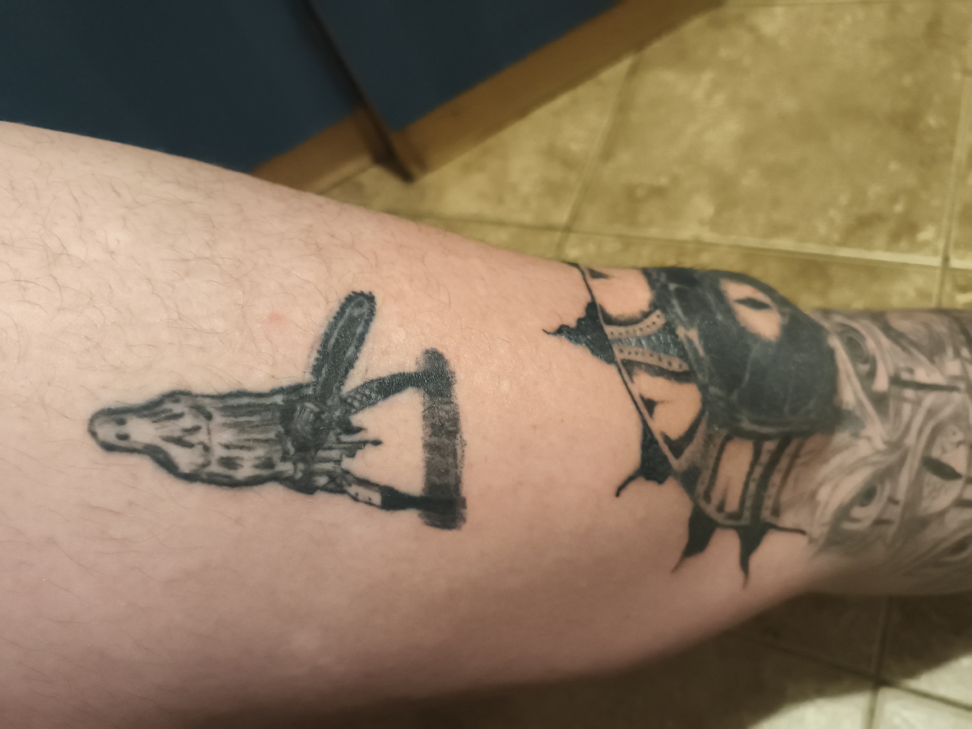 The image shows a portion of a person's leg with two distinct tattoos. On the left side of the frame, there is a black ink tattoo of what appears to be an alligator-like figure with skeletal features. To the right of this, closer to the knee, is a larger, more detailed black ink tattoo, with a design that is not fully discernible due to the angle but appears to incorporate geometric and possibly human-like elements. The skin is fair and has noticeable hair. The background includes a glimpse of a blue object, possibly part of furniture, and beige, tiled flooring. The location in Nowy Tomyśl, Poland is not discernible from the image.