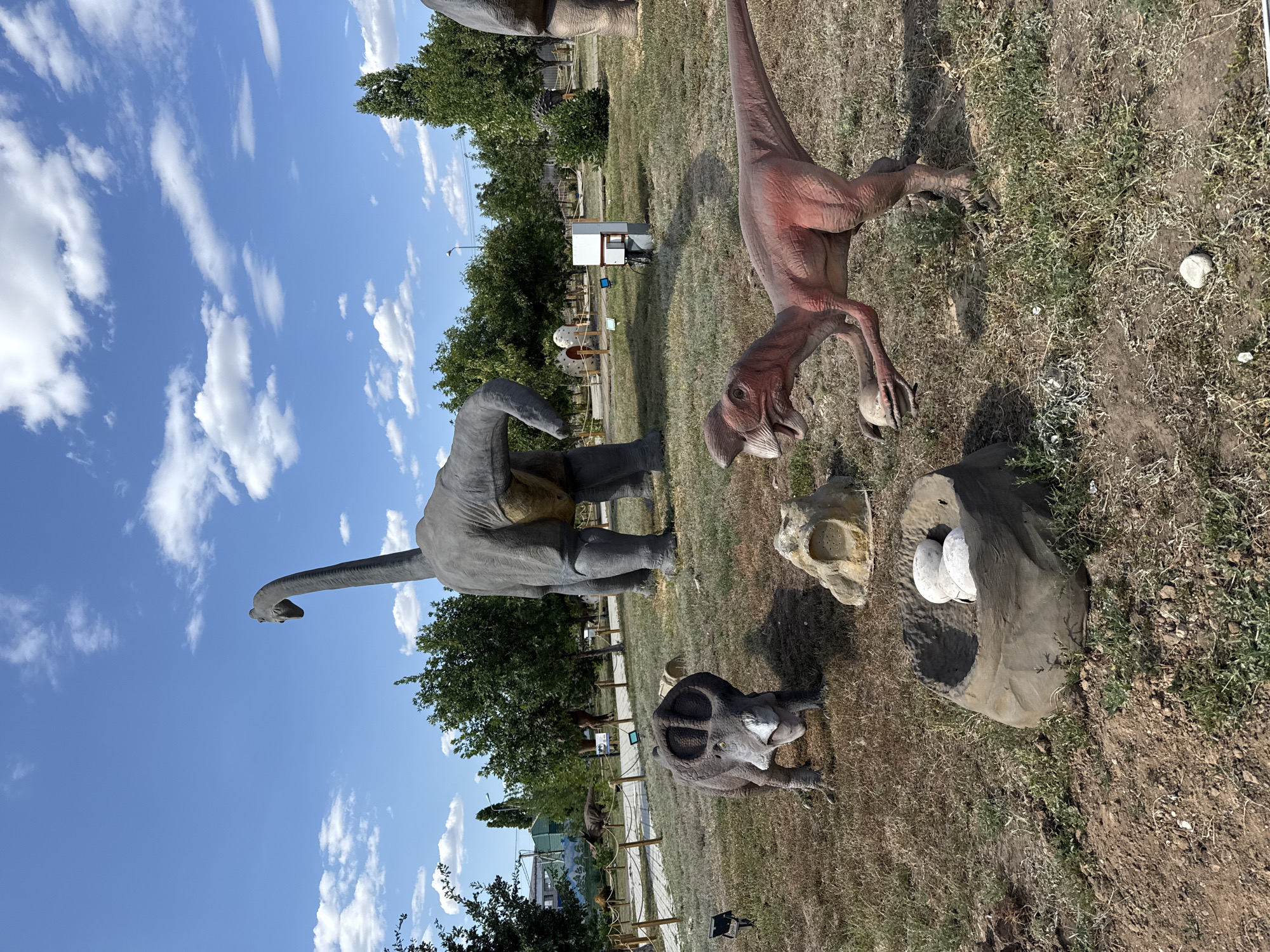 In the city of Sol-Iletsk, Russia, a dinosaur park is depicted under a bright blue sky with scattered white clouds. In the foreground, a model of a raptor dinosaur is shown holding a dinosaur egg in its clawed hand, standing near a nest containing three large, white eggs. To the left, a model of a Triceratops dinosaur faces forward, its horns prominent. In the background, a large Brachiosaurus model stands tall, its long neck reaching upwards. The scene is set in an outdoor area with dry grass, scattered rocks, and a few trees. A walking path and fencing are visible further in the background, along with other dinosaur models and park infrastructure.