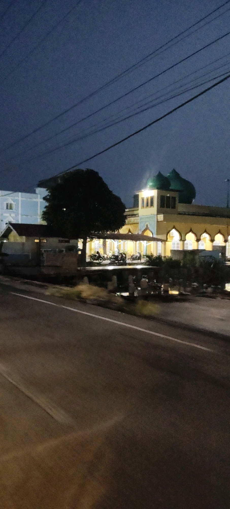 The image, taken at dusk or night in Sungai Pakning, Indonesia, depicts a street view with a prominent mosque. The mosque features green domes and a yellow facade with arched windows. In front of the mosque, there are several parked motorcycles. To the left of the mosque, a large, dark tree stands, partially obscuring a light blue building. The foreground shows a road with a white line, indicating it is likely a street or highway. Power lines are visible stretching across the dark sky. The overall atmosphere is dim, suggesting artificial lighting from the mosque and surrounding structures. There are no people clearly visible or identifiable in the image.