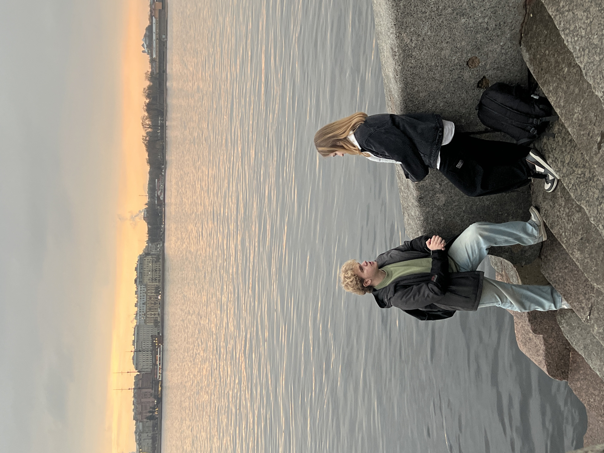 Two people are sitting on a granite embankment or low wall next to a body of water, likely a river, with a city skyline visible in the background. The scene is likely in Saint Petersburg, Russia.
One person, with long blonde hair, is wearing a dark jacket over a light shirt, and dark pants. A black backpack sits next to this individual. The other person has curly blonde hair, a light green shirt, a dark jacket, and light blue jeans.
The city skyline consists of buildings of various heights and architectural styles, with some trees interspersed. The water surface shows subtle ripples, reflecting the light from the sky, which appears to be undergoing a sunset. The granite embankment is gray and rough in texture.