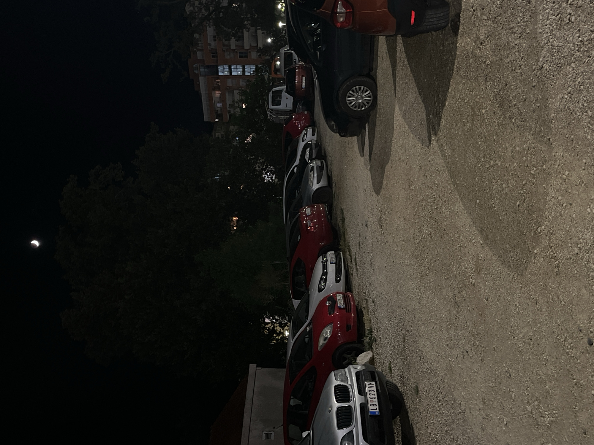 The image depicts a nighttime scene in a parking lot in Budva, Montenegro. The parking lot is surfaced with gravel and is partially illuminated by artificial lights from nearby buildings. Several cars are parked in the lot, including a silver BMW X5 with license plate "LB 023 VM", a red Toyota Yaris, a white Volkswagen Golf, a red car, and a dark colored car in the foreground with a flattened tire. In the background, a multi-story building with illuminated windows is visible, as well as trees and a sliver of the moon in the dark sky. There are no people visible in the image. The overall impression is that of a quiet, dimly lit parking area.