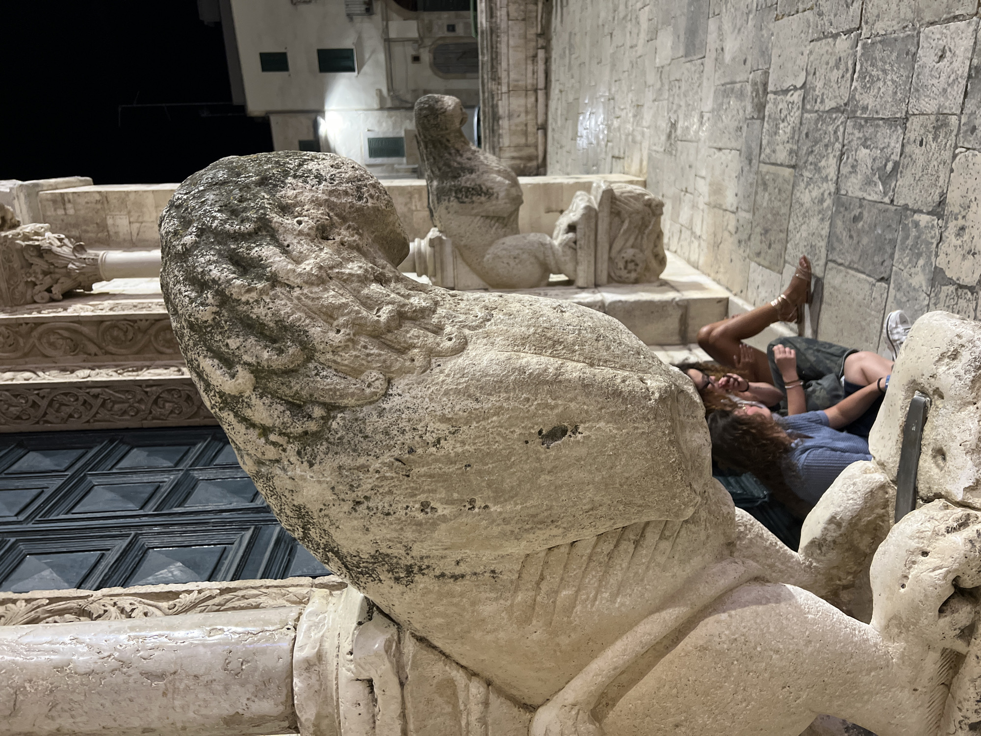 The image shows a nighttime view of the exterior of the Basilica of San Nicola Pellegrino in Bitonto, Italy, focusing on a large, weathered stone lion sculpture in the foreground. Two people are seated beneath the sculpture, partially obscured by its bulk. One person, wearing a blue shirt, is looking towards the left, while another person, wearing a camouflage-patterned top, is looking towards the right. A third person's legs, wearing a gold sandal, are visible to the right. The pavement is made of stone slabs. In the background, other architectural elements of the basilica and buildings in the town are visible. A smaller lion sculpture is also present further in the background.