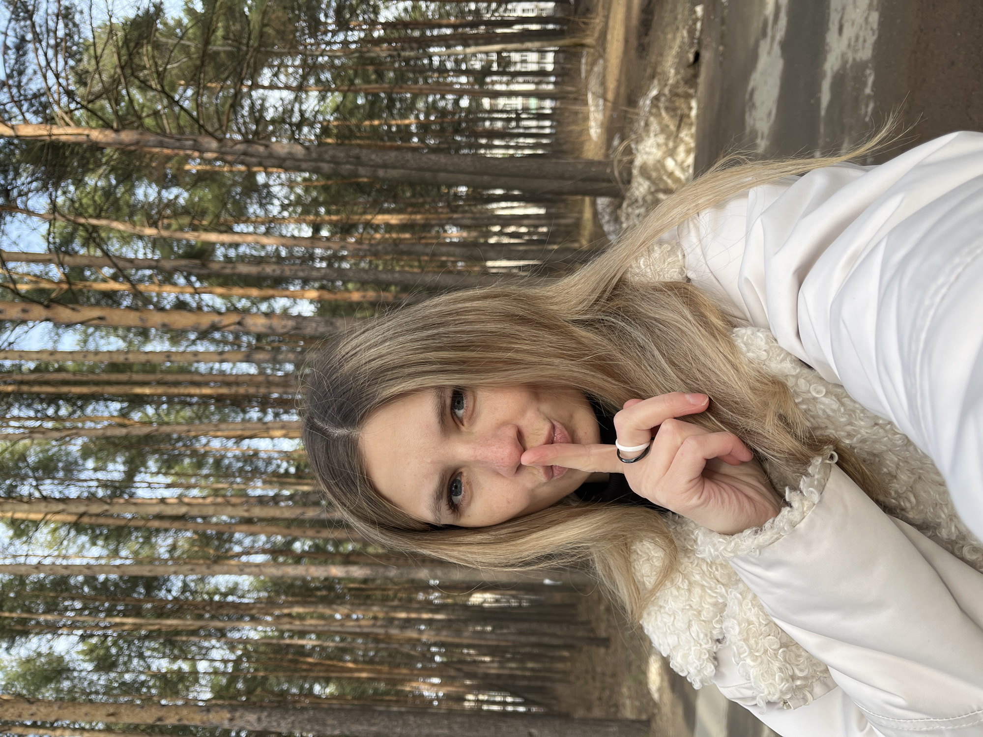 A Caucasian woman with blonde hair is photographed outdoors. She is wearing a white jacket with a shearling collar. She is holding a finger to her lips. She has two rings on her right ring finger, one black and one white. The background is a forest of tall, thin trees. The location is in Chaikovsky, Russia.