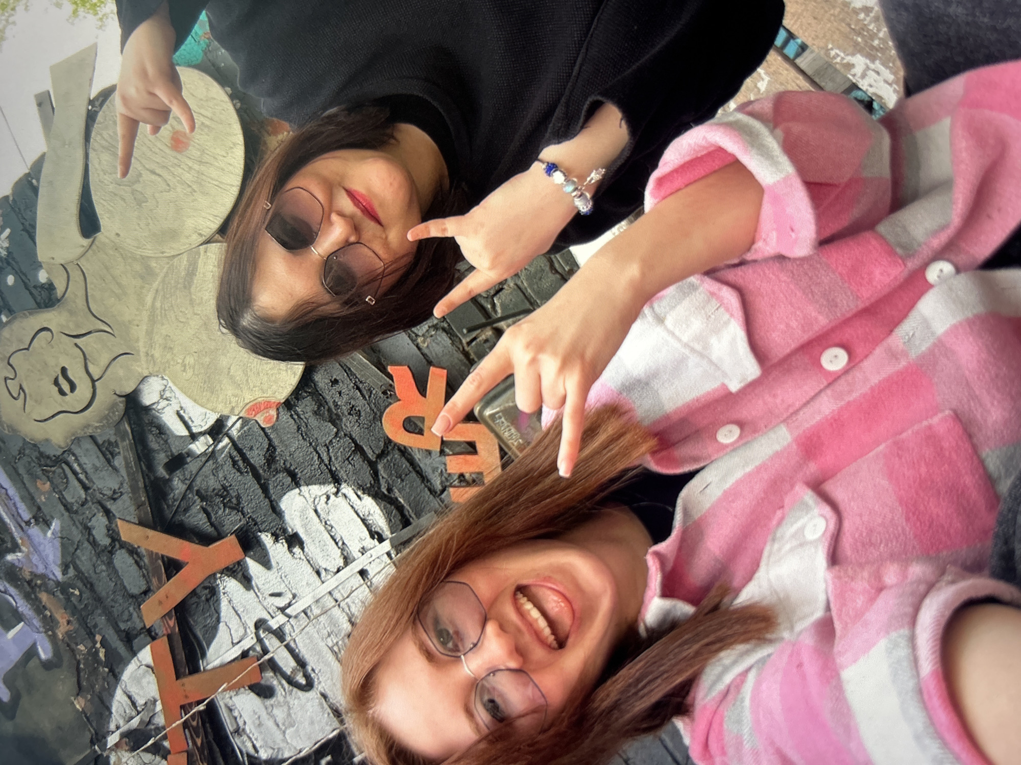 The image shows a close-up selfie of two young women. The woman in the foreground has long, reddish-brown hair and is wearing glasses and a pink and white checkered shirt. She is smiling with her mouth open and her tongue visible. The woman in the background has dark brown hair and is wearing sunglasses and a black top. She is also smiling and holding up two fingers on each hand in a rock-and-roll gesture.
The background appears to be an outdoor setting with a textured surface, possibly brick or concrete, and some artistic elements. There are some metal or wooden cutouts visible in the background, one of which seems to depict a stylized character. Orange letters are also visible, forming part of some text. The lighting suggests it is daytime. The overall mood of the image is fun and energetic, with both women appearing to be in good spirits.