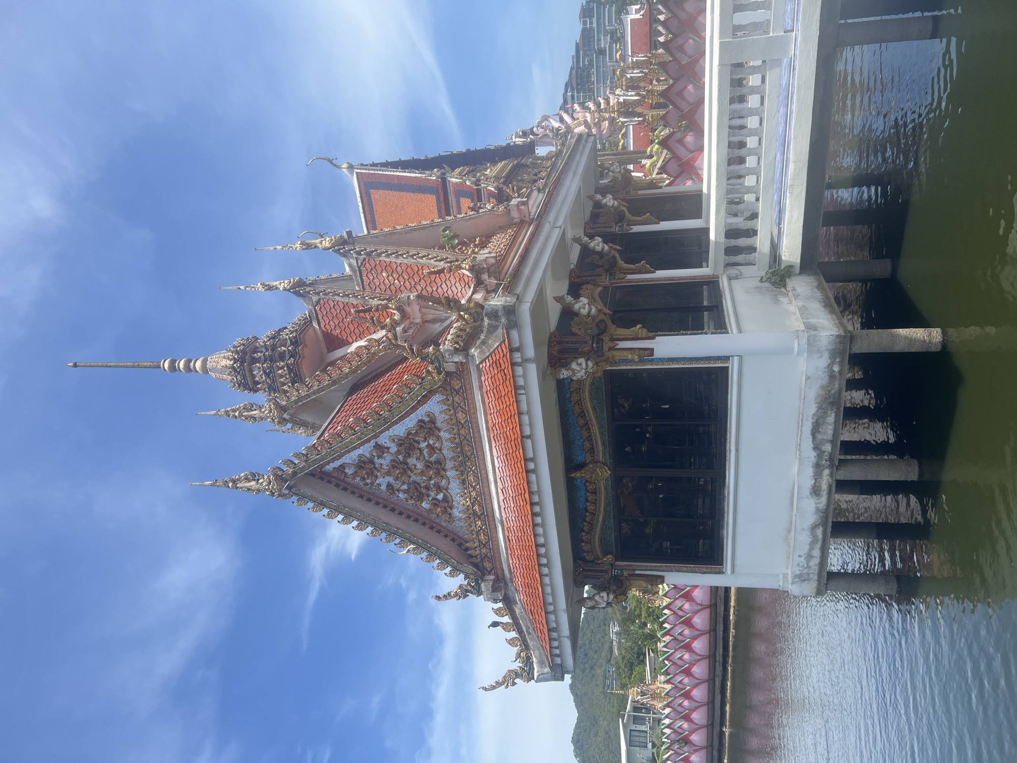 The image captures a stunning view of a Thai Buddhist temple situated over the water in Ko Samui District, Thailand. The temple's architecture is ornate, featuring multi-tiered roofs with intricate golden carvings and vibrant red tiles. The main structure rests on stilts above the water, connected by a white walkway with a decorative railing. Surrounding the temple is a body of water, likely a pond or a calm bay, reflecting the blue sky and clouds. In the background, lush green hills and some modern buildings can be seen, suggesting a developed area. The scene is bathed in bright sunlight, indicating a clear day.