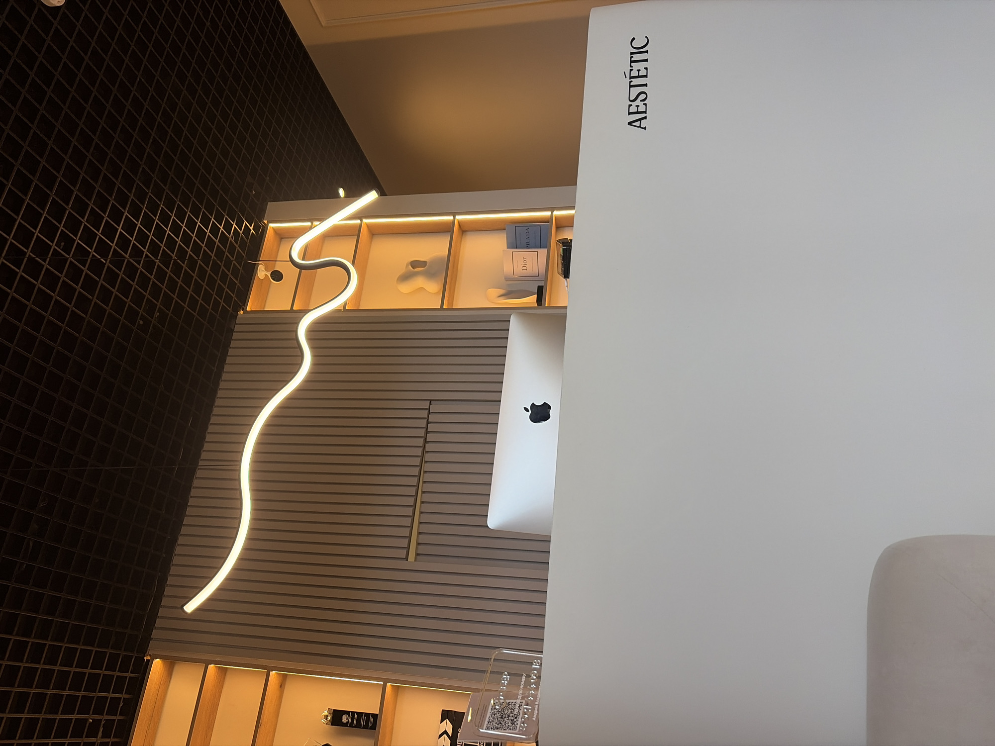 This image captures a modern and elegant interior space, likely a commercial establishment such as a beauty clinic, salon, or design studio, located in Saint Petersburg, Russia. The perspective is upward-looking, showcasing contemporary design elements and warm lighting.
To the right, a prominent white wall or partition displays the word "AESTÉTIC" vertically in a clean, black sans-serif typeface. Behind this partition, the back of an Apple iMac computer is visible, featuring its distinct black Apple logo. Partially seen in the bottom right is a light-colored, upholstered chair or sofa, suggesting a waiting or consultation area.
The central portion of the image features a horizontal slatted wall made of light-toned wood or panels. Above this, several backlit display niches or shelves showcase various decorative items, including abstract white sculptures and luxury brand books, with titles "Dior" and "PRADA" clearly legible. A long, serpentine-shaped LED light fixture, emitting a soft, warm glow, traverses the upper part of the room, acting as a striking design feature.
On the far left, a wall is clad in dark, small square tiles, creating a textured grid pattern. Near the bottom, a transparent plaque with a QR code and unreadable text, possibly Braille, is partially visible. No people are present, and the scene is static, emphasizing the pristine and sophisticated aesthetic of the environment. The overall impression is one of modern luxury and meticulous design.