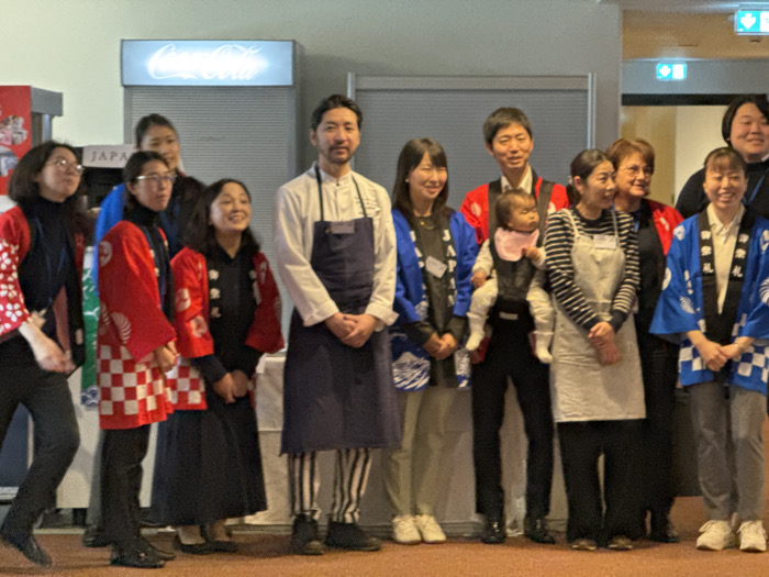 An indoor photograph in Strasbourg, France, captures approximately 14 individuals, predominantly adults of East Asian descent, posing in what appears to be a public event space. The environment features a reddish-brown carpeted floor and light-colored walls. In the background, an illuminated Coca-Cola sign, a partially visible "JAPA" sign, and green emergency exit indicators are present. The individuals are arranged in an informal line, mostly facing the camera with smiles.
The group's attire includes multiple traditional Japanese happi coats in varying designs: some are red with white checkered patterns or white wave motifs, while others are blue with white wave patterns. A central male figure, identifiable as a chef, wears a white chef's jacket, a dark apron, and black-and-white striped pants. To his right, another man carries an infant in a dark front-facing baby carrier; the baby wears a pink bib and light-colored clothing. Adjacent to them, a woman wears a striped long-sleeve shirt and a light-colored apron. One Caucasian woman is also visible among the group, wearing a red happi coat. The collective presentation suggests a Japanese cultural or culinary event.