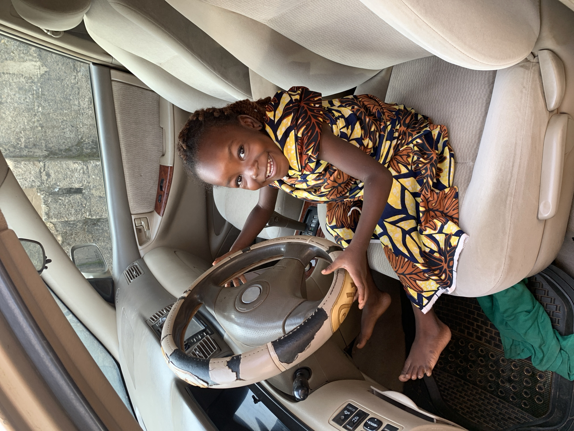 A young Black girl is sitting in the driver's seat of a car, smiling at the camera. The car's interior appears worn, with a cracked steering wheel cover and faded upholstery. The girl is wearing a colorful, patterned dress and has her hands on the steering wheel. Her bare feet are visible. A green cloth is on the floor mat. The background shows a rough, concrete wall outside the car window. This scene is located in the Sabon Tasha area of Kaduna, Nigeria, based on the context provided.