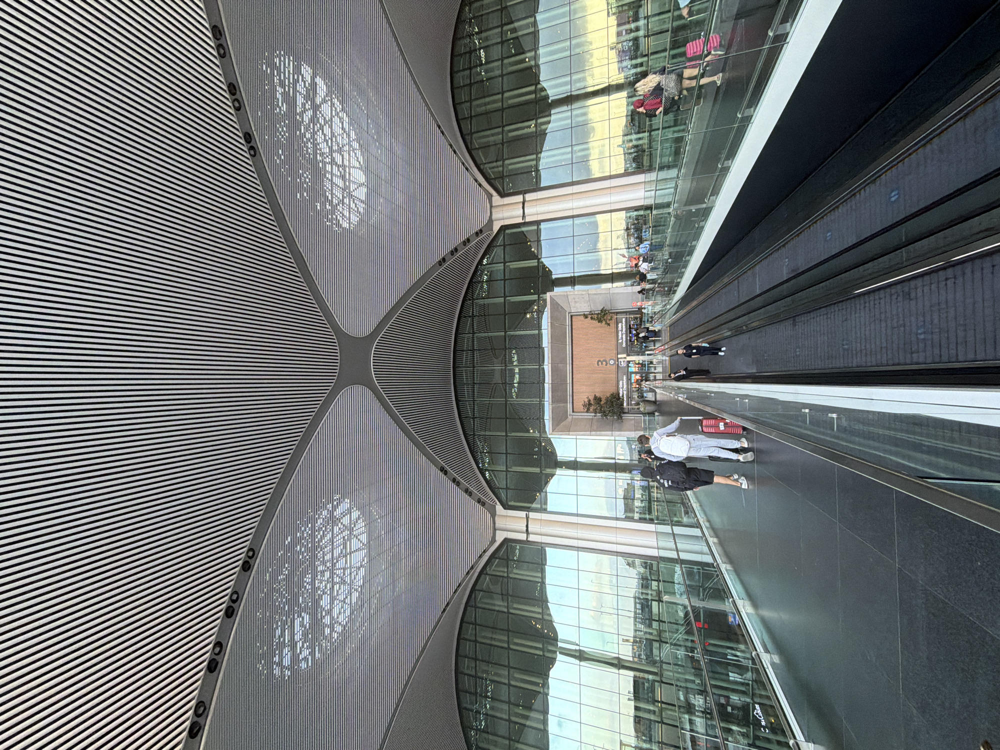 The image depicts the interior of a modern airport terminal in Arnavutköy, Turkey. The ceiling features an intricate, ribbed design with large, curved architectural elements. Floor-to-ceiling glass walls offer a view of the exterior, which appears to be a bright, possibly late afternoon or early morning scene with reflections of the sky and buildings.
In the foreground, a walkway runs parallel to two escalators. On the walkway, three people are visible. Two individuals, a male and a female, walk towards the background, the male carrying a black backpack and the female pulling a red suitcase. Another person, a male, walks ahead of them on the walkway.
On the upper level, a glass-enclosed walkway is populated with several people. Some are walking, while others appear to be standing. A woman with blonde hair is seen pulling a pink suitcase.
The escalators in the foreground are empty and moving. The overall environment suggests a busy transportation hub with clean, contemporary architecture. Signs are visible in the background, including one that says "Iç Hatlar Only" and "Domestic Departures," indicating this is likely a domestic terminal area. A small tree is planted near an entrance.