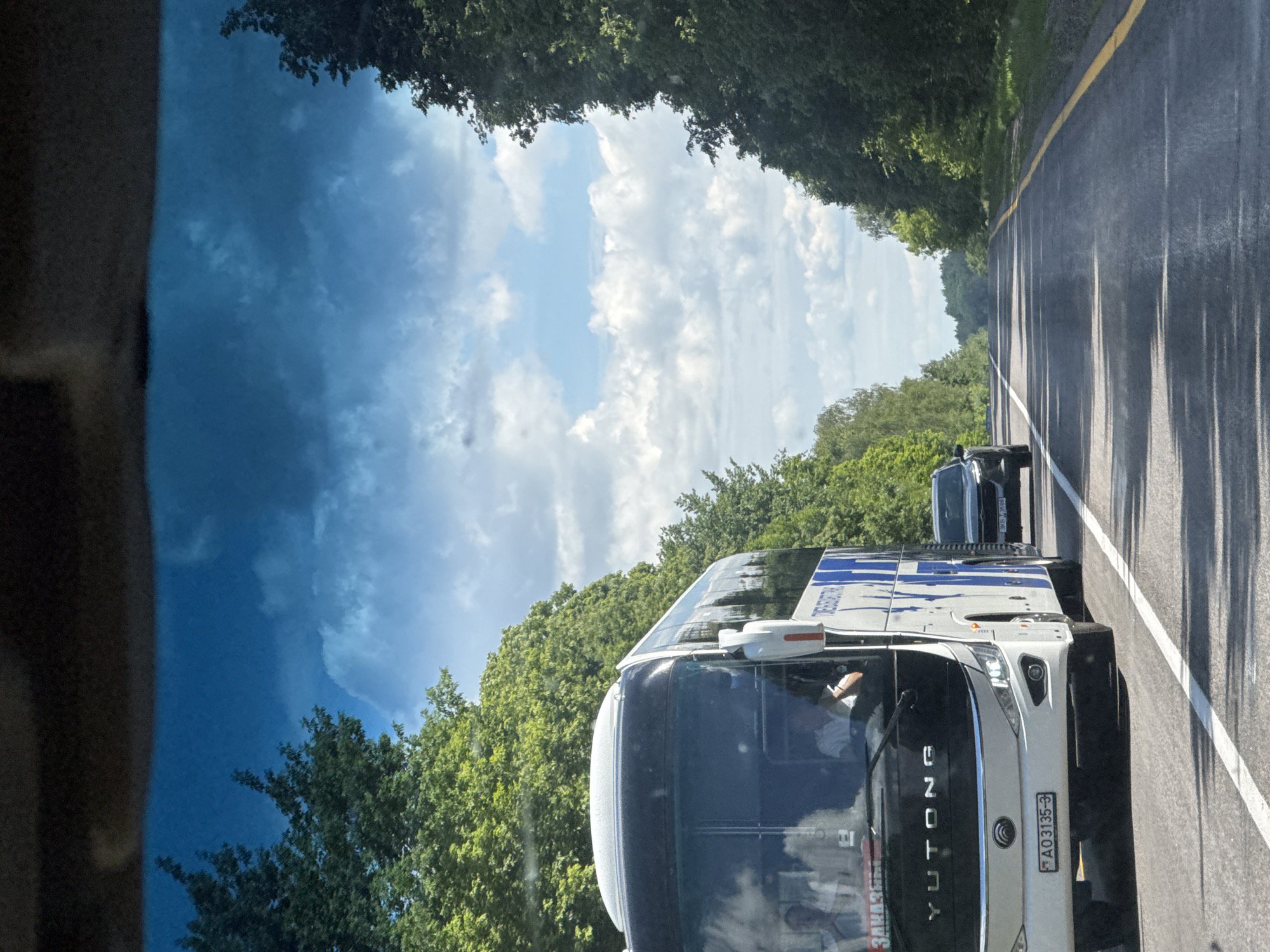 This is a video taken from inside a vehicle, possibly a car or a bus, driving on a road lined with lush green trees. The road curves to the right, with a white line marking the edge and a yellow line in the center, indicating a two-way highway. Sunlight casts long shadows on the asphalt, suggesting it is daytime, likely afternoon, and the weather is bright and sunny with scattered clouds in a blue sky.

The primary subjects are a white tour bus with blue stripes and the text "YUTONG" visible on its front, and a smaller dark vehicle behind it. The bus has a license plate that appears to read "FA03135-3". The perspective suggests the camera is positioned slightly above and to the left of the bus's front. Inside the bus, a person is visible in the driver's seat.

The scene conveys a sense of travel and movement through a scenic, wooded area, likely in Belarus given the context of Роўкавіцкі сельскі Савет. The bright sky and shadows indicate pleasant weather, and the overall mood is peaceful and typical of a road trip. There is no discernible activity or event beyond the vehicles driving along the road.