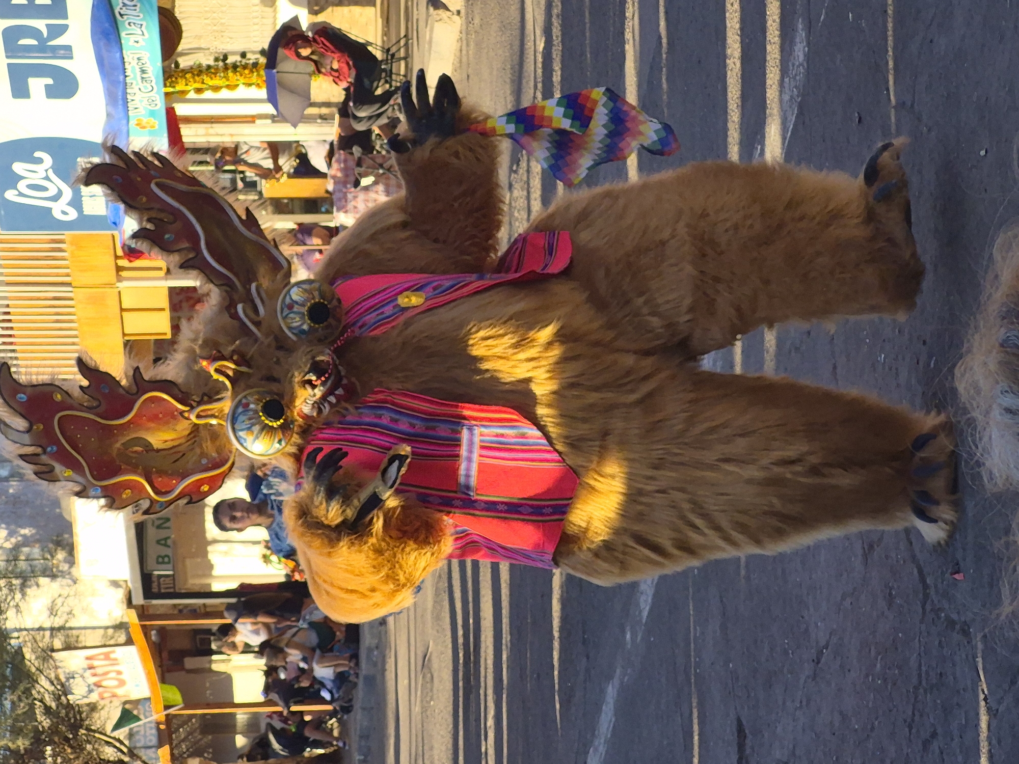 In Pozo Almonte, Chile, a person in a furry, brown costume with large, antler-like headdresses and exaggerated facial features is walking on a paved street. The costume has a colorful, striped vest and the person is holding a small, multi-colored cloth in their right hand. In the background, there are buildings with signage, some people watching from the side, and a few parked vehicles. The environment appears to be an outdoor street scene, possibly during a festival or parade, with sunlight casting shadows on the ground.