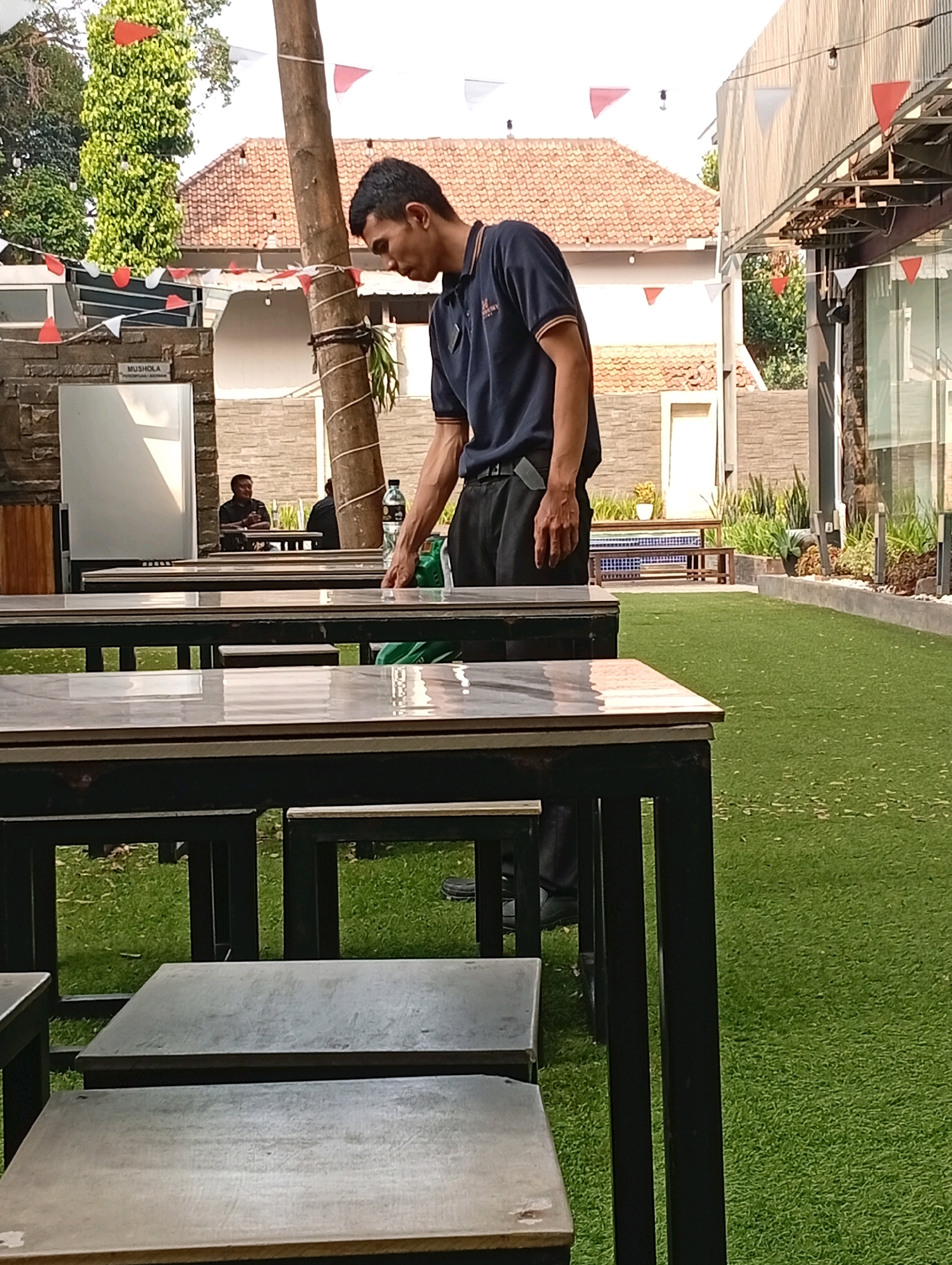 The image captures an outdoor seating area, likely a cafe or restaurant, with artificial turf covering the ground. In the foreground, several tables with dark frames and light-colored tops are arranged. A man, appearing to be an employee, is visible in the mid-ground, wearing a dark blue polo shirt and dark trousers. He is bending over and appears to be attending to something on a table. In the background, there is a building with a tiled roof, and flags are strung across the scene, suggesting a festive occasion. The overall environment appears to be a public space in Binong, Indonesia, designed for outdoor dining and relaxation.